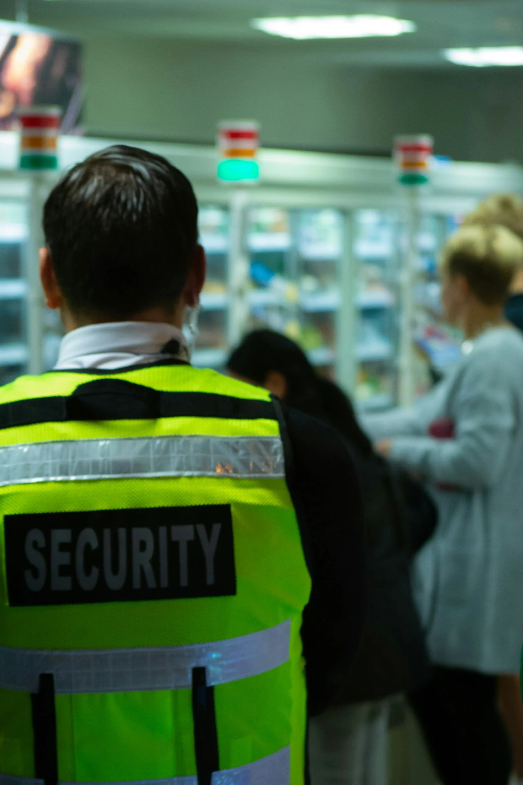 Security guard in fluorescent yellow vest with 'SECURITY' label, facing the crowd at an indoor location, likely an airport or train station.