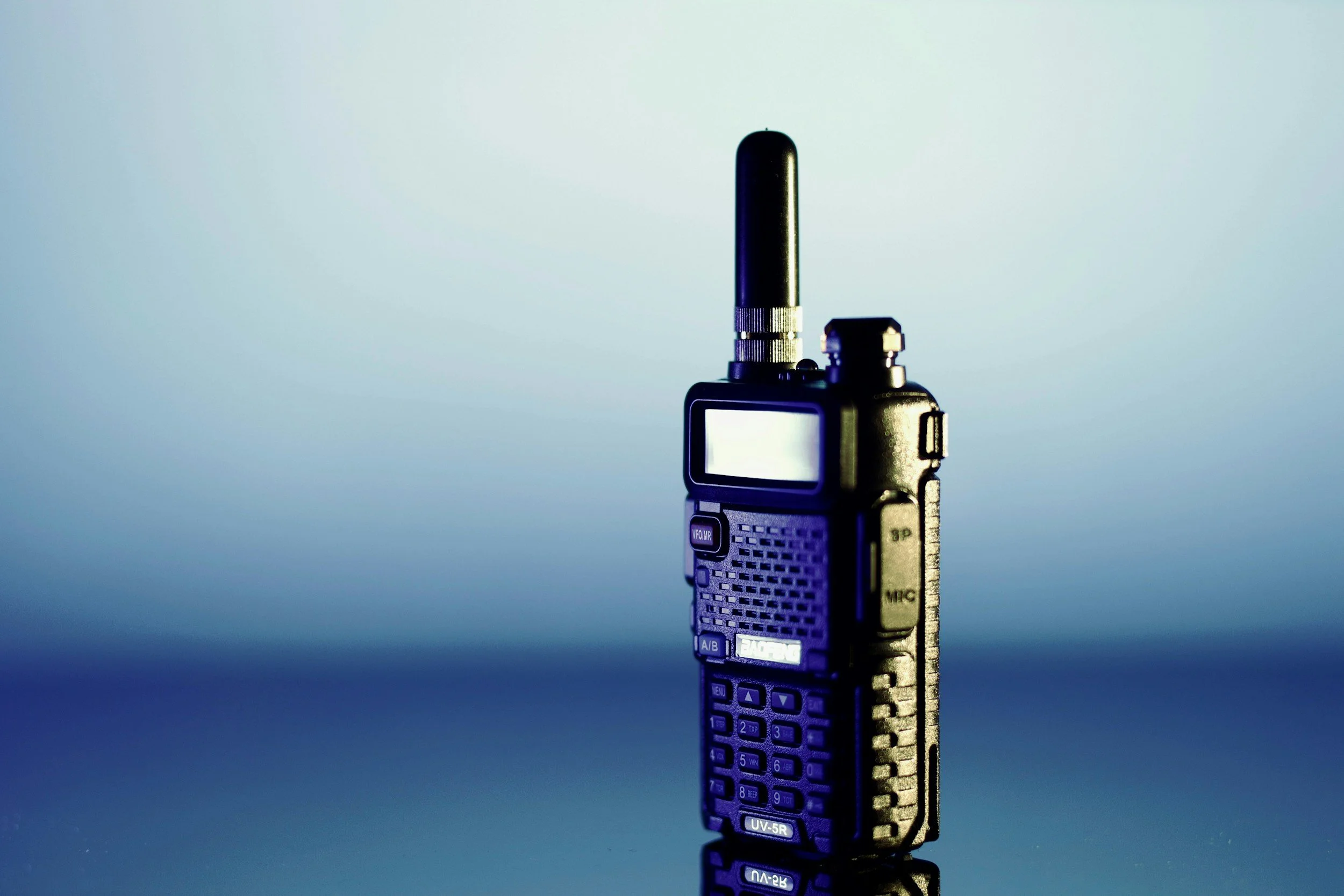 A black two-way radio walkie-talkie with an antenna standing upright on a reflective surface with a gradient blue background.