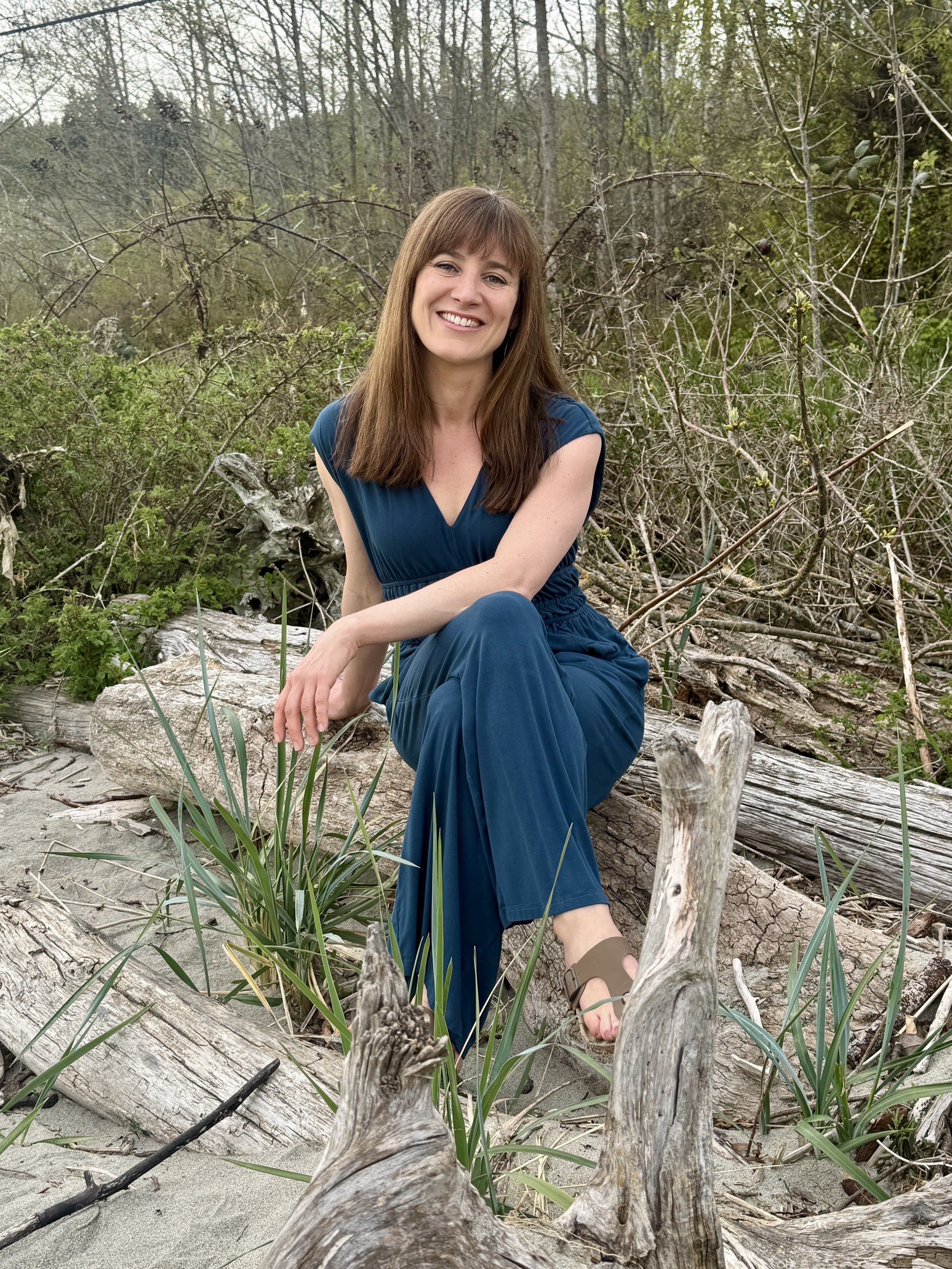 A woman with long brown hair in a dark blue dress sitting on driftwood and grass in a wooded outdoor setting, smiling at the camera.