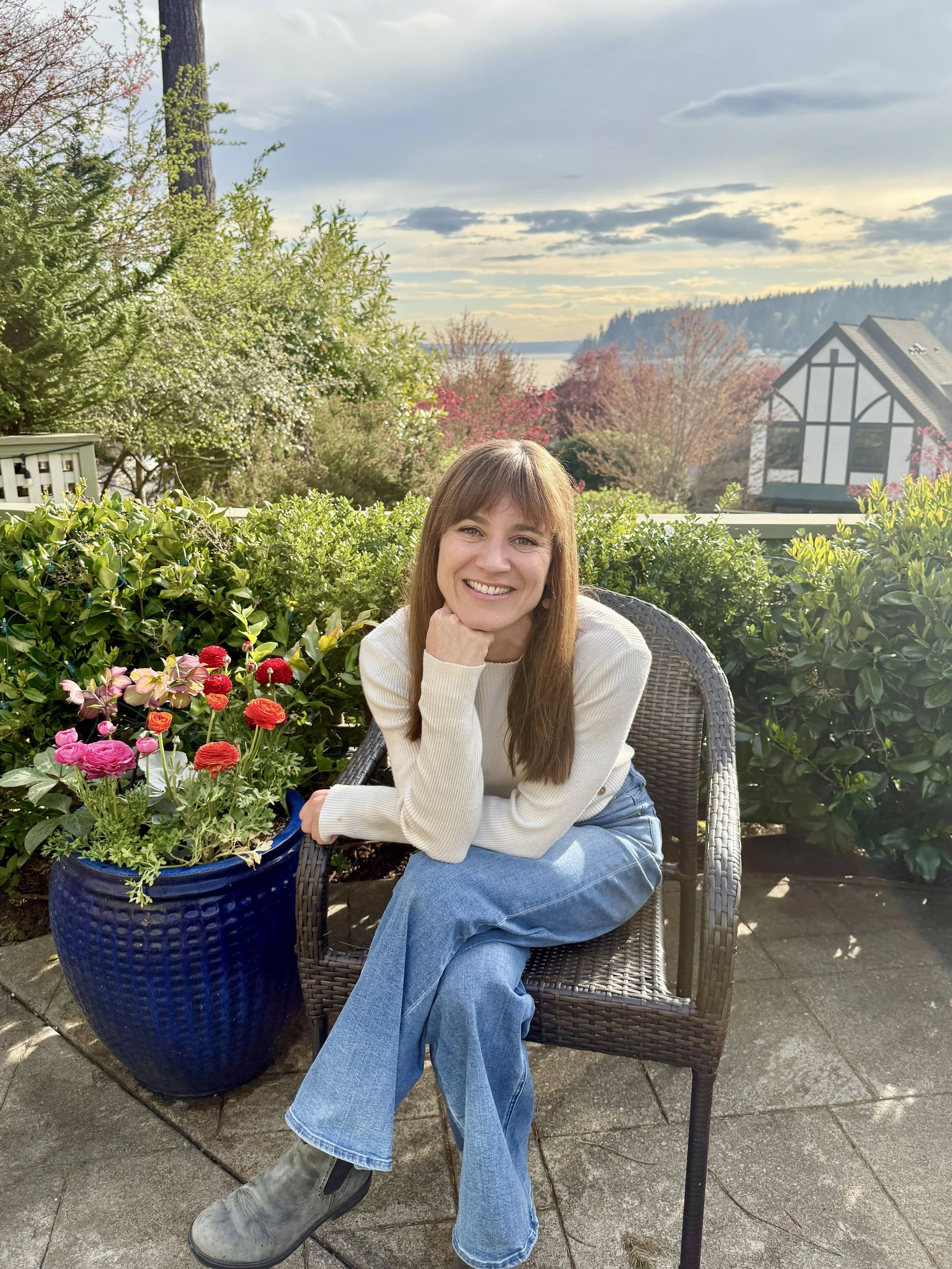 A woman smiling and sitting in a wicker chair on a patio, with a large blue flower pot filled with pink and red flowers beside her. She is wearing a white sweater, blue jeans, and gray boots, with a garden and trees in the background under a partly cloudy sky.
