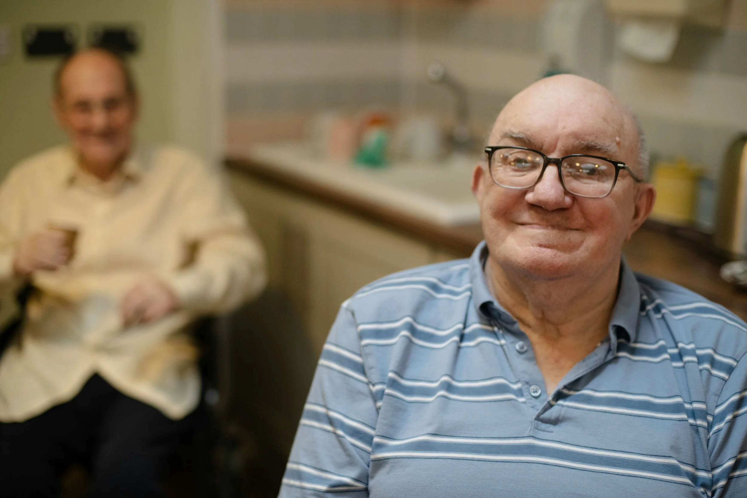 A smiling bald man with glasses wearing a striped blue polo shirt in the foreground with a woman in a yellow blouse in the background, both in a kitchen setting.