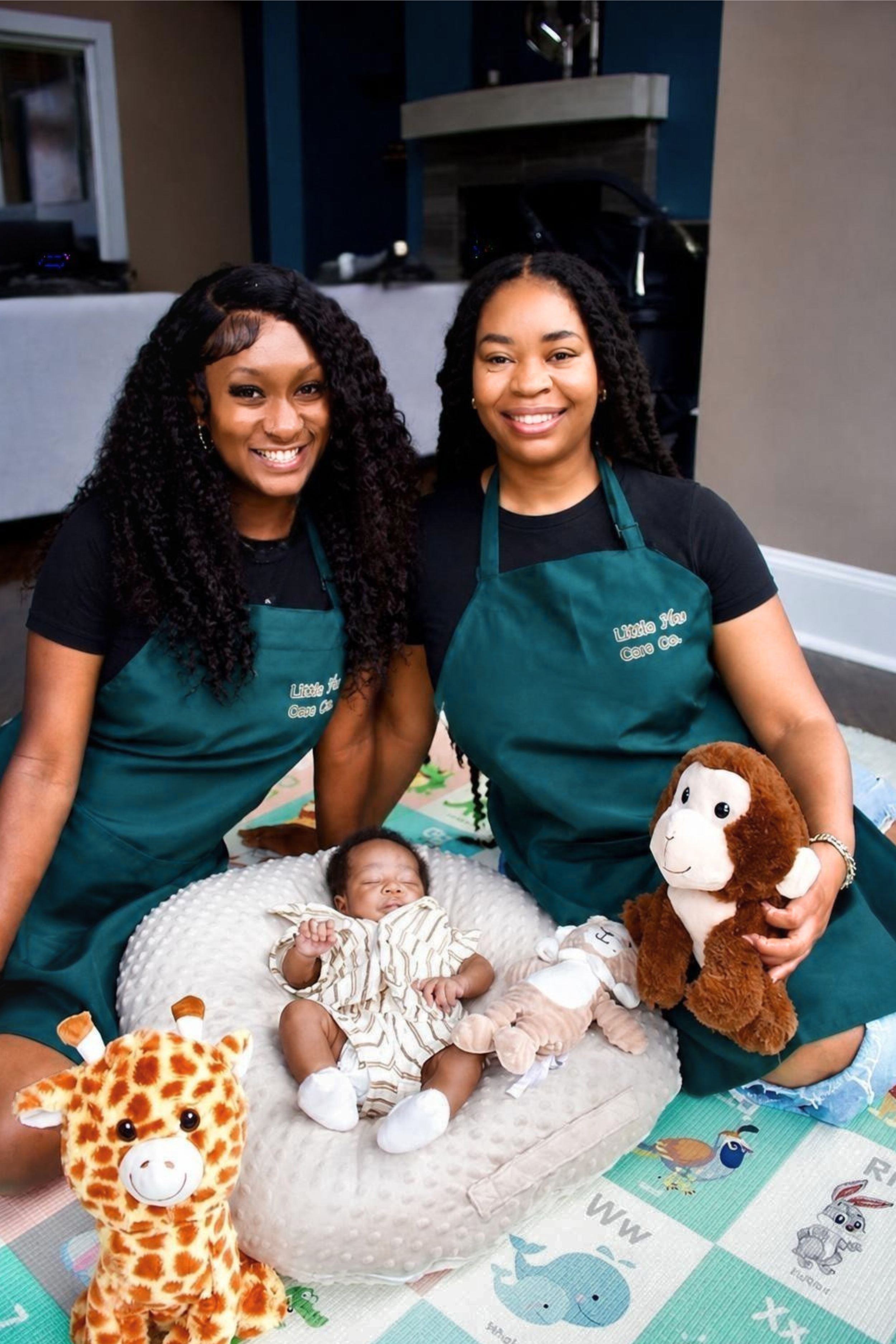 Two nurturing women wearing forest green aprons with the words "Little Plus One Co." pose with a baby on a cushioned pillow, holding stuffed animals and plush toys. They are indoors, standing on a colorful play mat with animal illustrations.