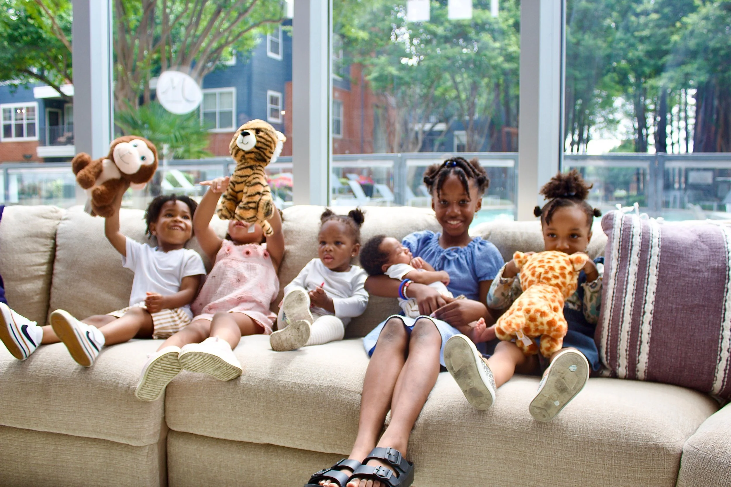 Six children sitting on a beige couch in a sunlit room with large windows, holding and playing with plush animal toys, including lions and a tiger, with a view of trees outside.