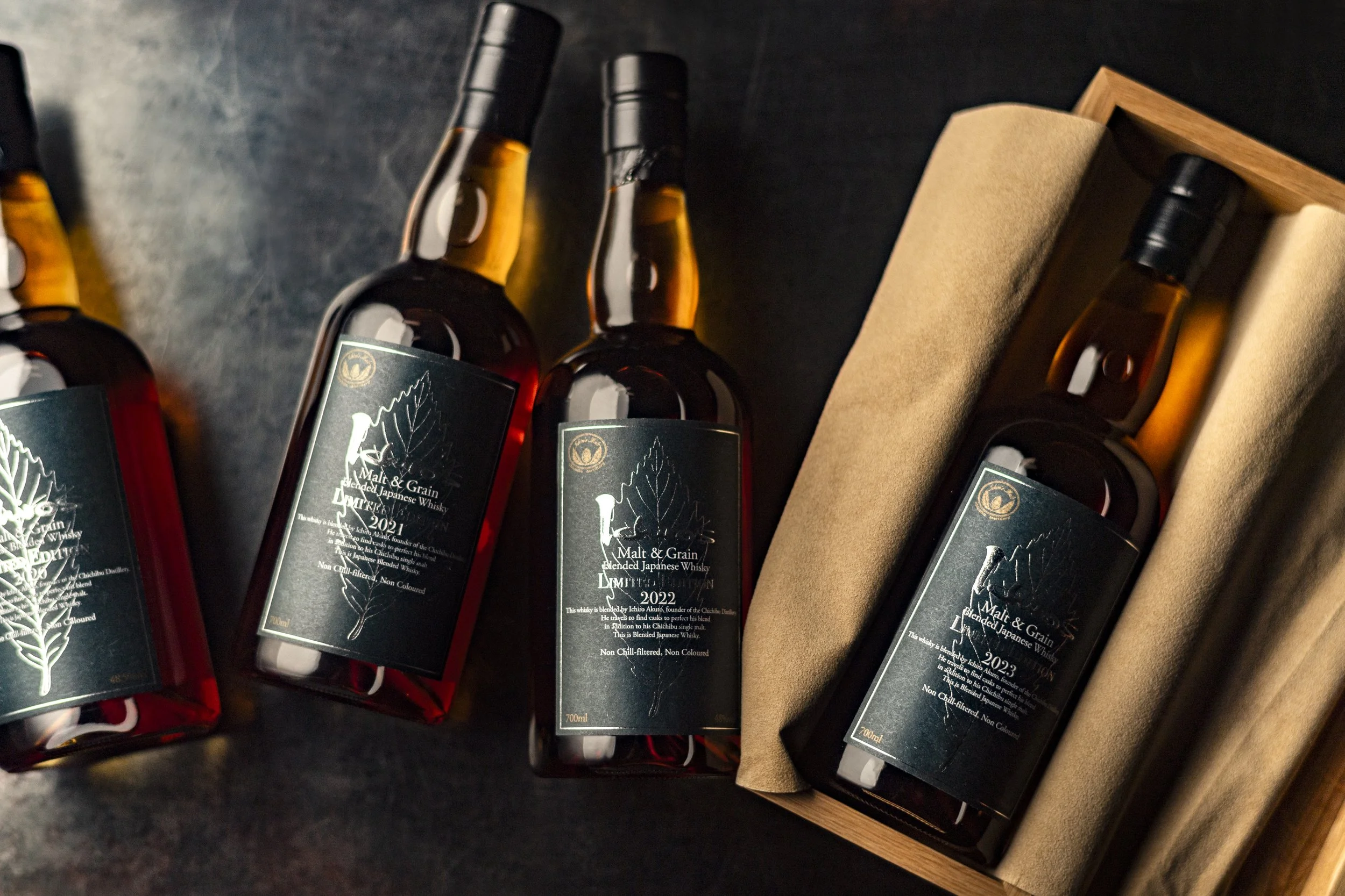 A set of three bottles of blended Japanese whisky with black labels and gold caps, one of which is in a wooden box with a beige inner lining.