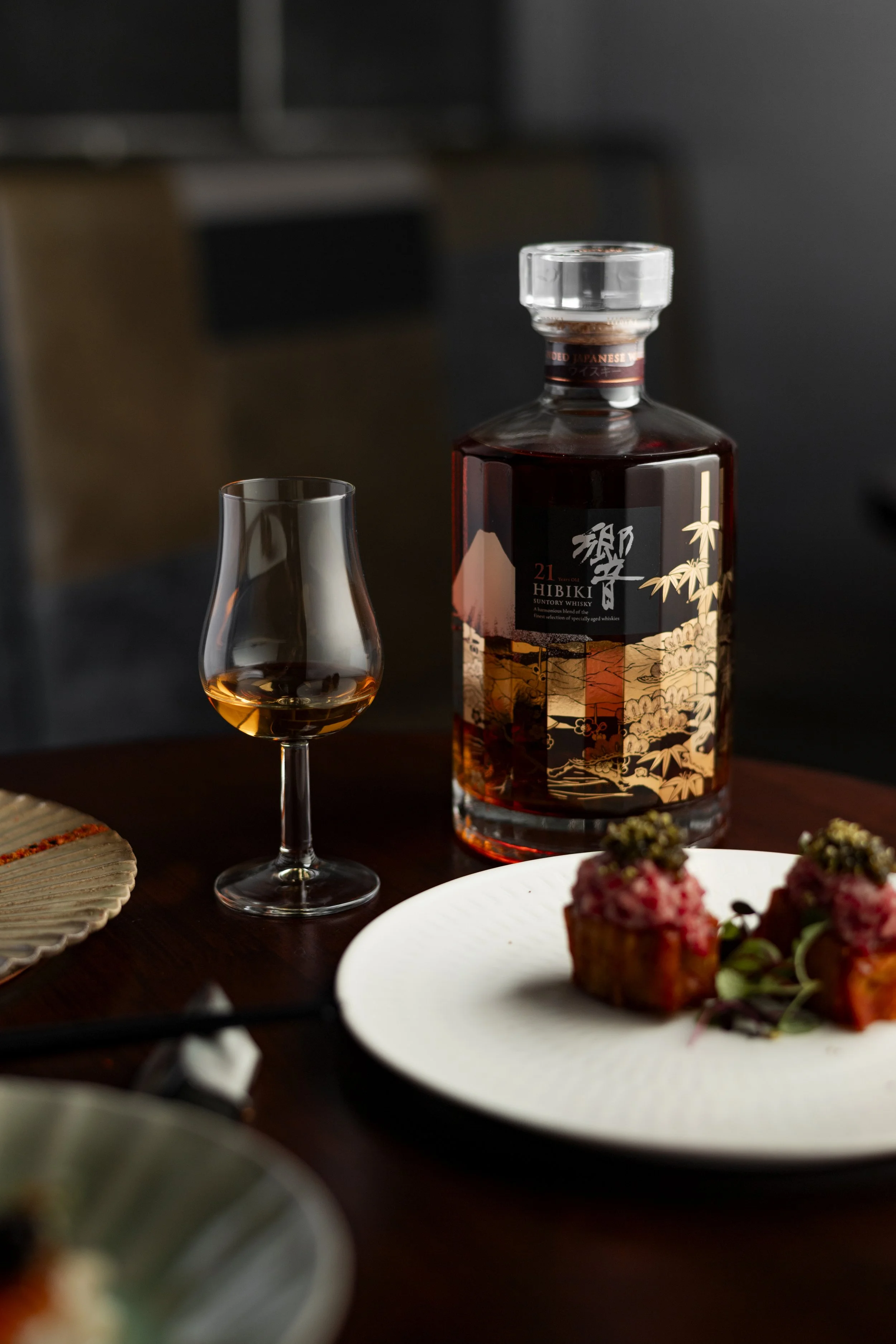 A bottle of Japanese Hibiki whiskey, a glass with a small amount of whiskey, and a white plate with two desserts topped with berries on a dark wooden table.