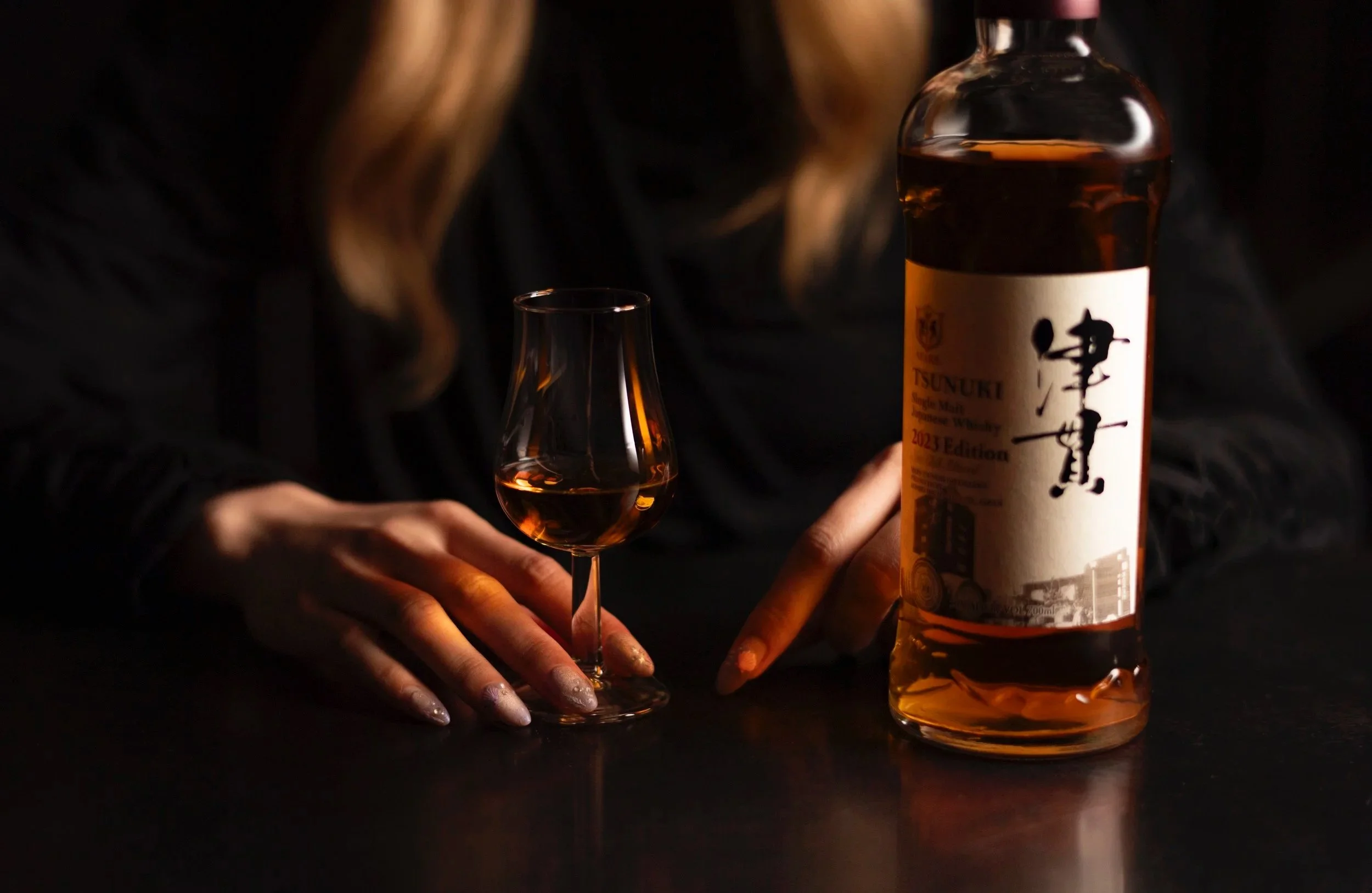 A person with long blonde hair holds a glass of amber-colored whiskey next to a bottle of Tsunuki single malt Japanese whisky on a dark surface.