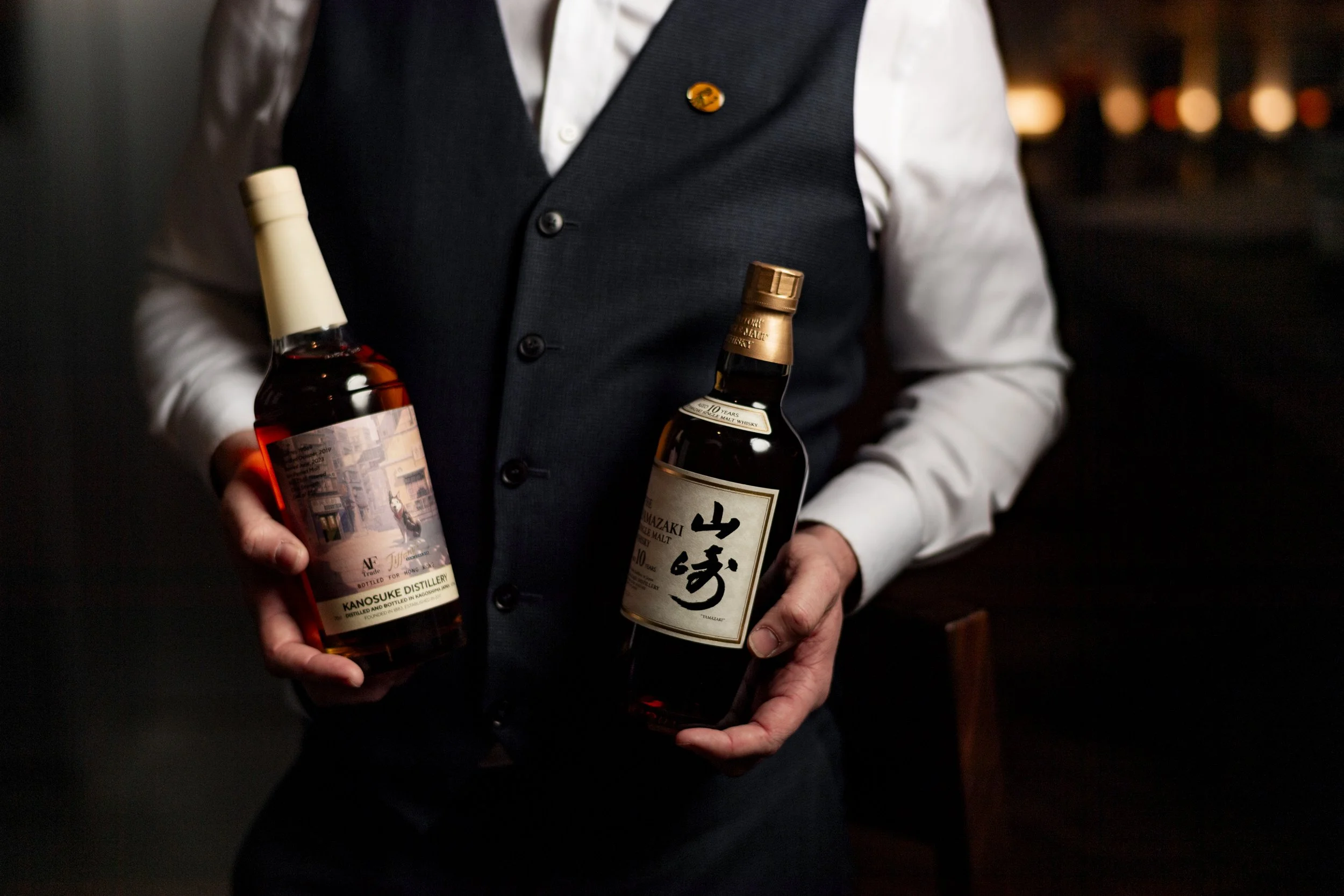 Person holding a bottle of whiskey and a bottle of Japanese whisky in a dimly lit setting.