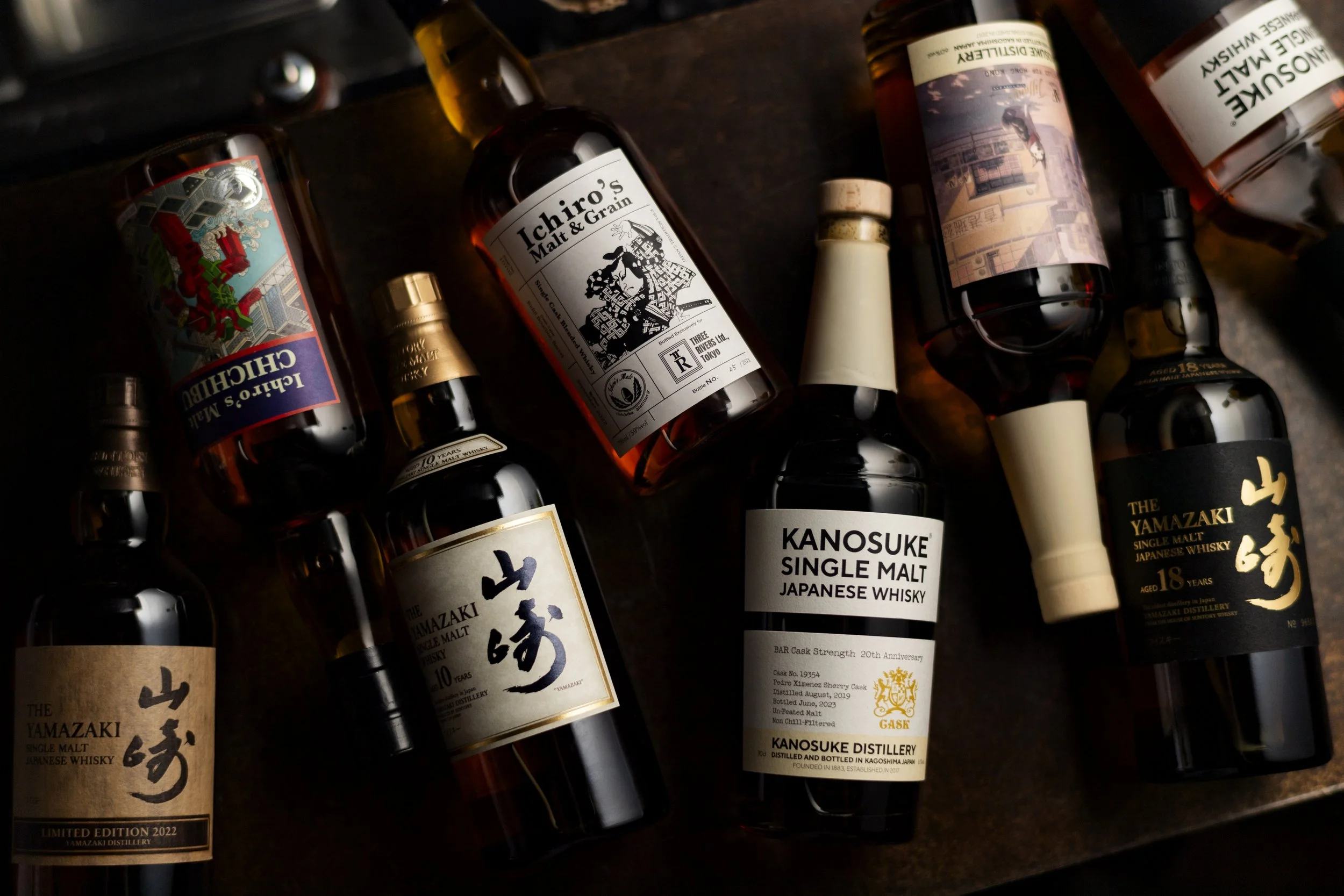 Various bottles of Japanese whisky on a dark surface, including Yamazaki, Kanosuke, Ichiro's Malt, and Hakushu.