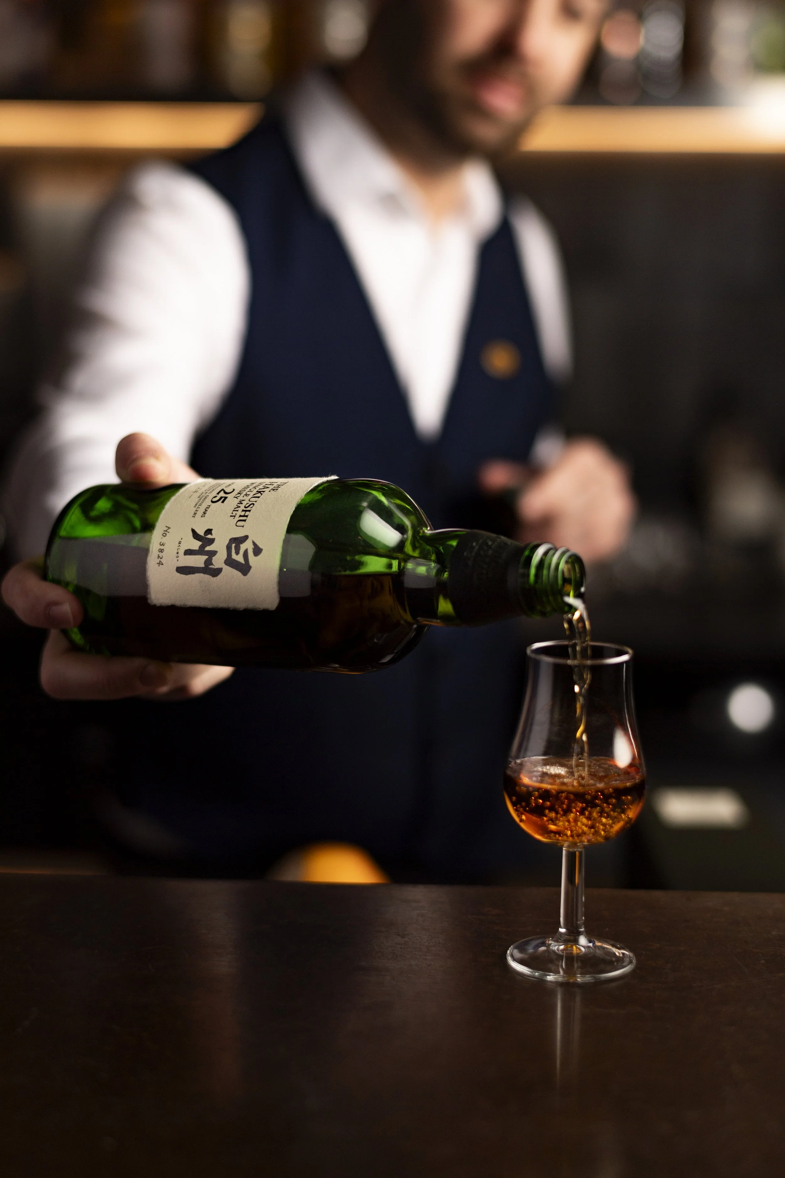 A bartender pouring amber-colored liquor from a green bottle into a glass on a dark bar counter.