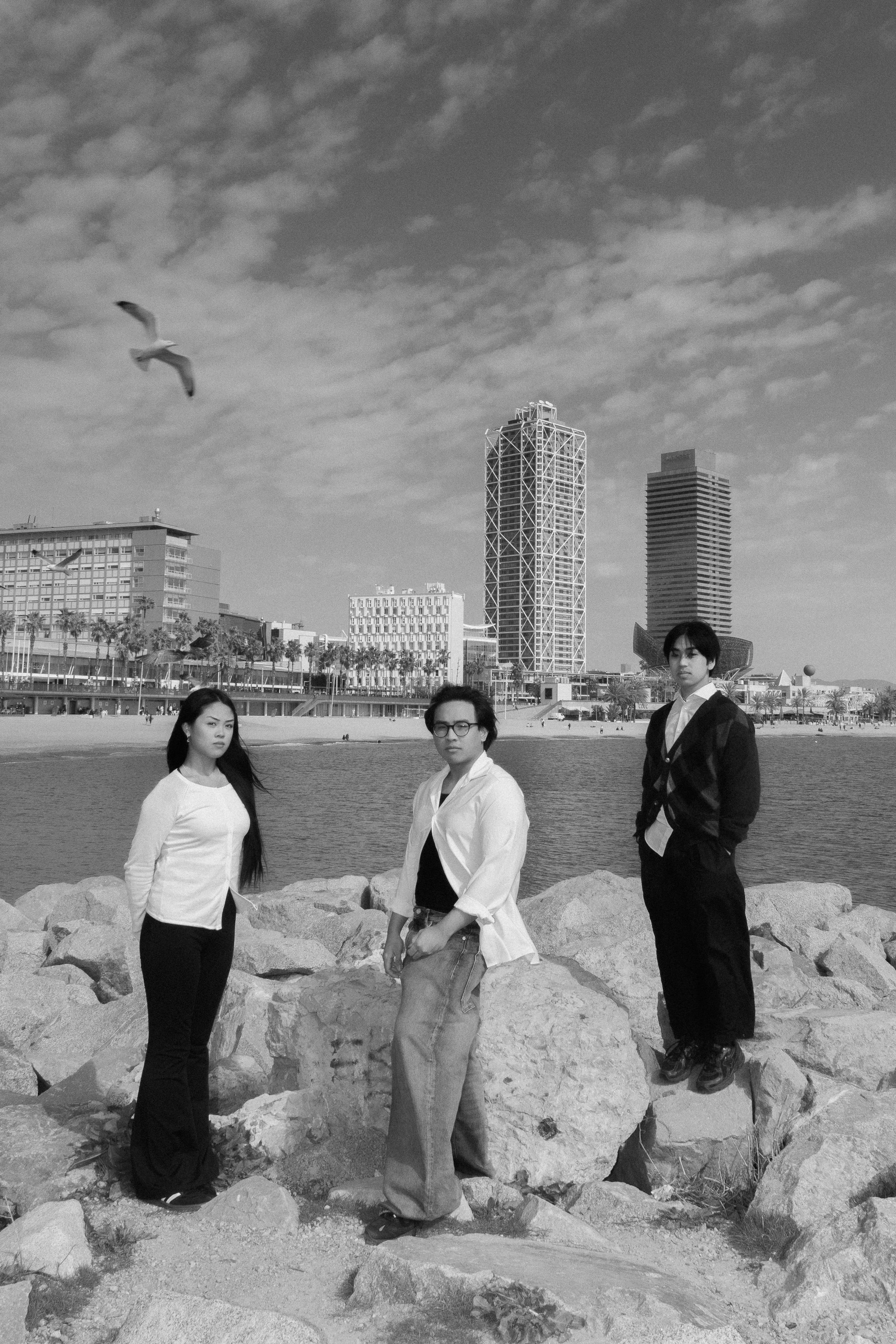 Three young adults standing on rocks near a waterfront with city buildings in the background, under a sky with scattered clouds, one woman and two men posing for a photo.