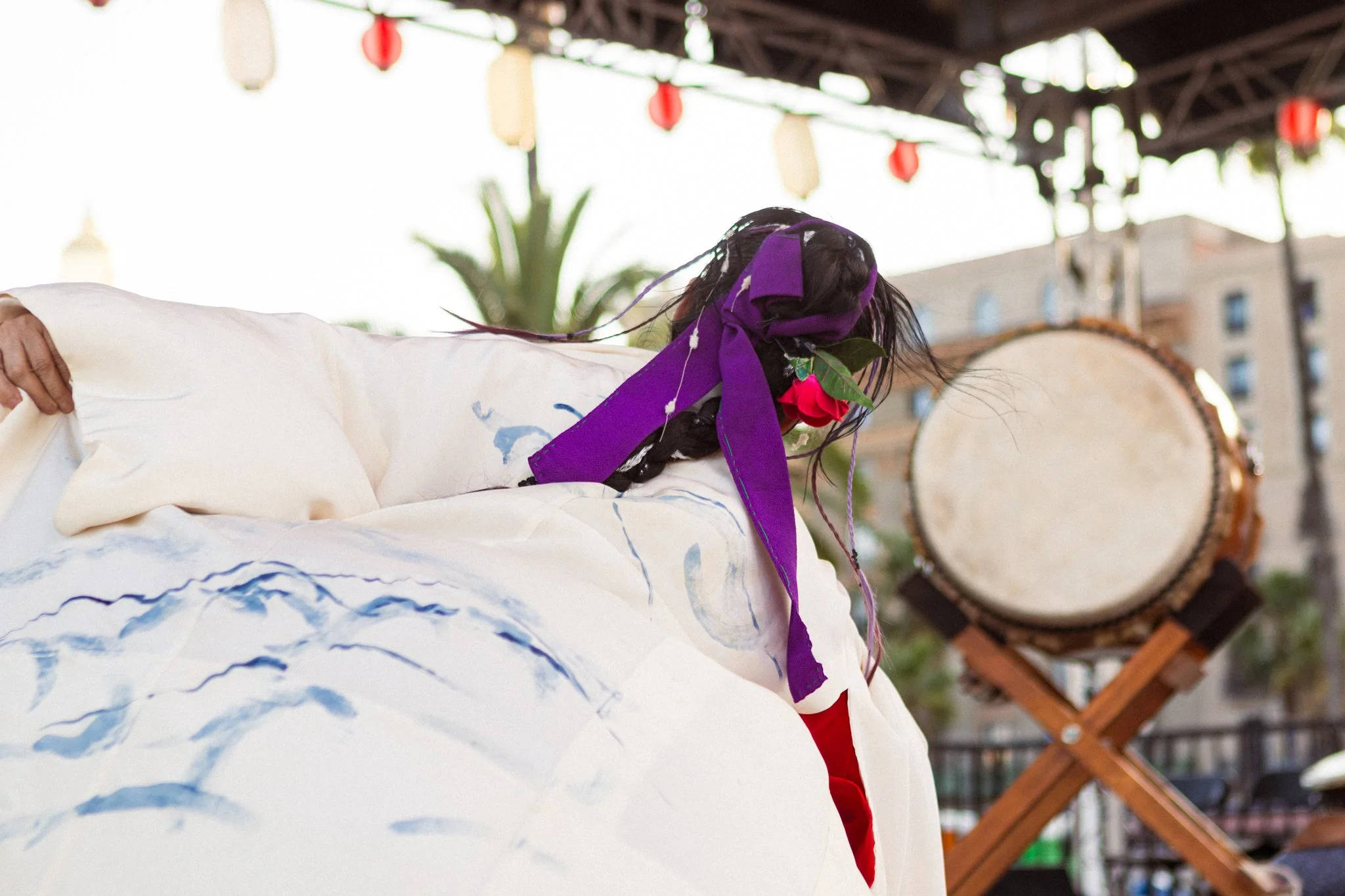 A person with long black hair wearing a purple headband and red lipstick is lying on a large, decorated white fabric with blue patterns. The person is holding the fabric with one hand, and there is a large white drum in the background. There are colorful lanterns hanging overhead, and buildings and trees are visible in the background.
