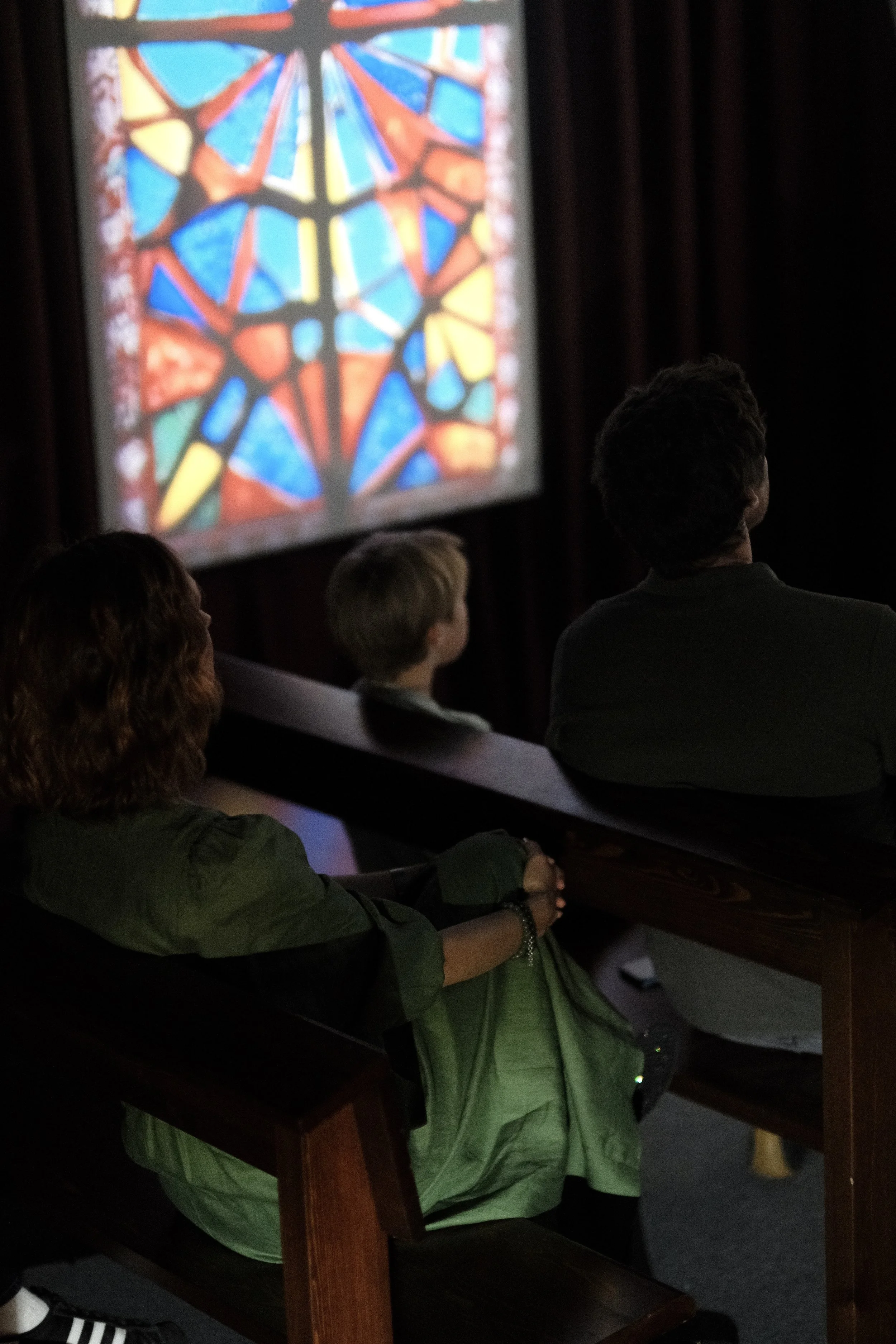 People sitting on wooden pews in a dark room, watching a colorful stained glass window projection.
