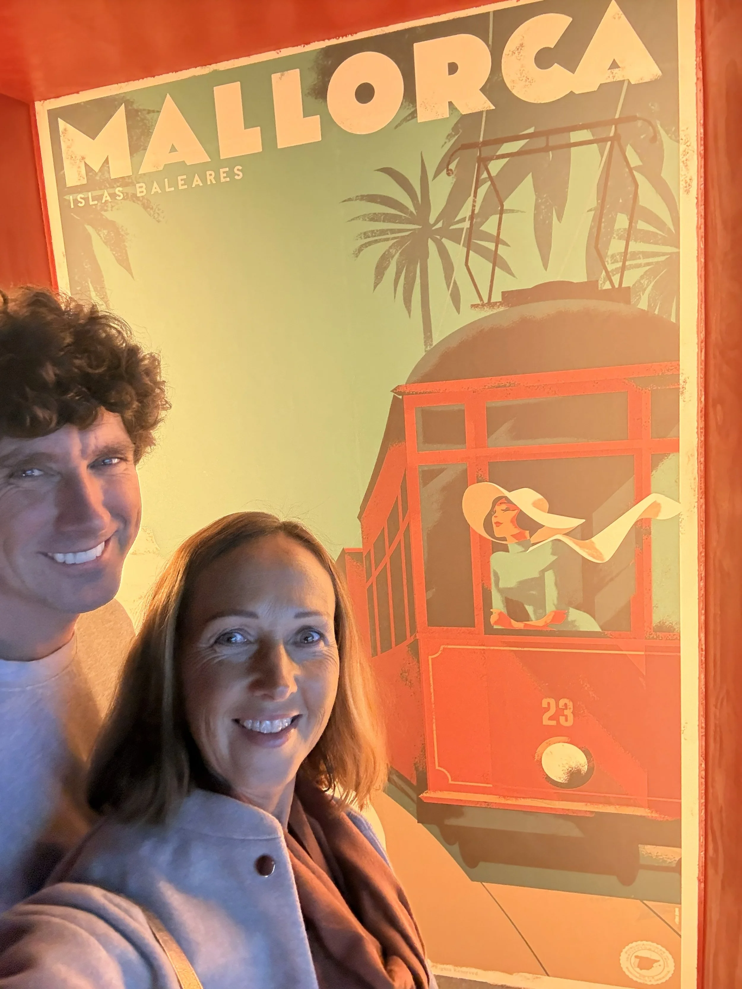 People smiling in front of a colorful vintage poster of Mallorca, Islas Baleares, featuring a woman in a sunhat in a tram with palm trees in the background.