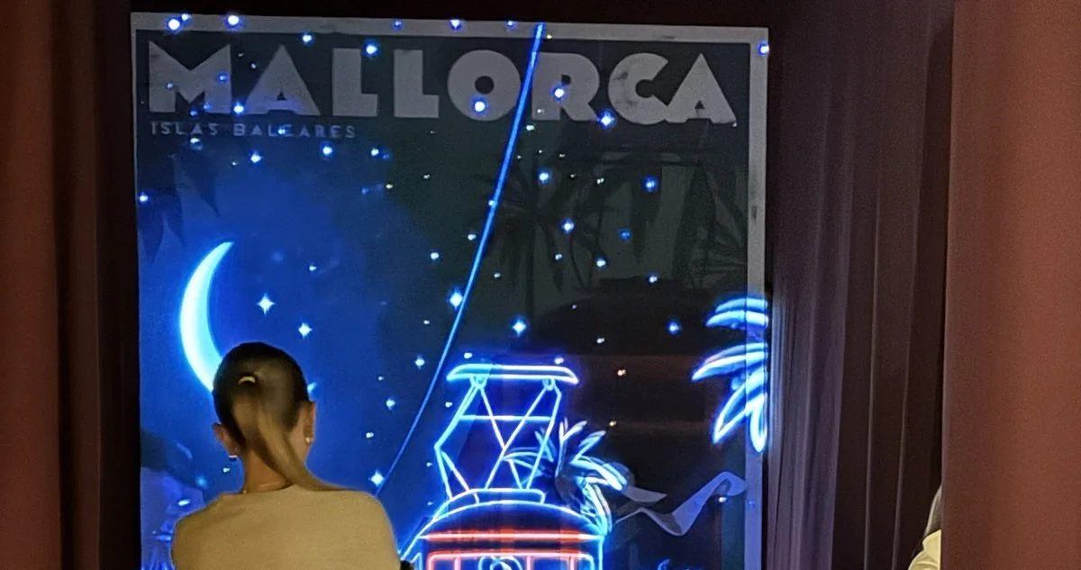 Person looking at a neon sign window display with the word 'Mallorca', images of a crescent moon, stars, a swing, and palm trees at night.