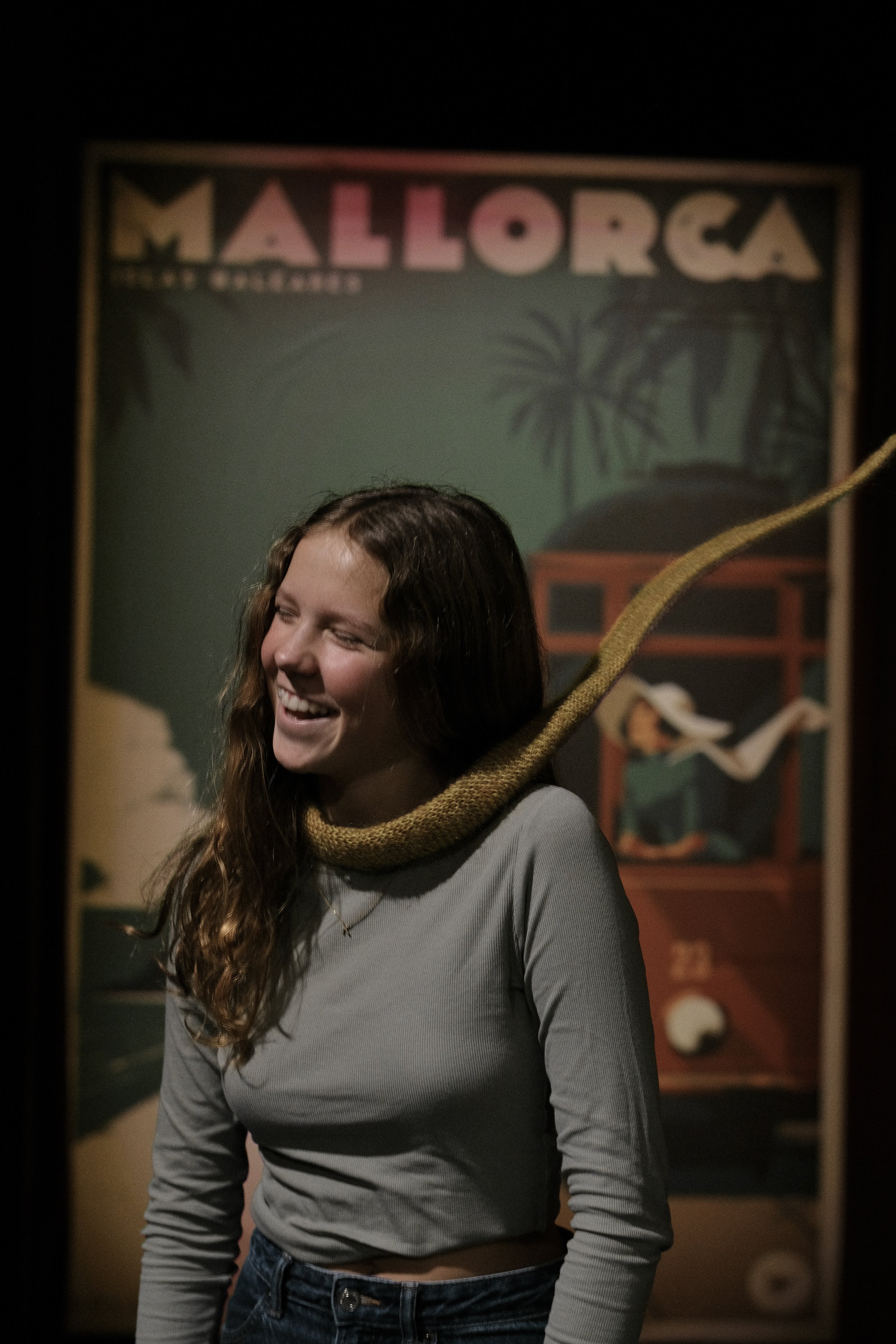 Young woman with curly hair, smiling, wearing a gray long-sleeve shirt and jeans, with a large yellow snake around her neck, standing in front of a 'Mallorca' poster in a dark room.