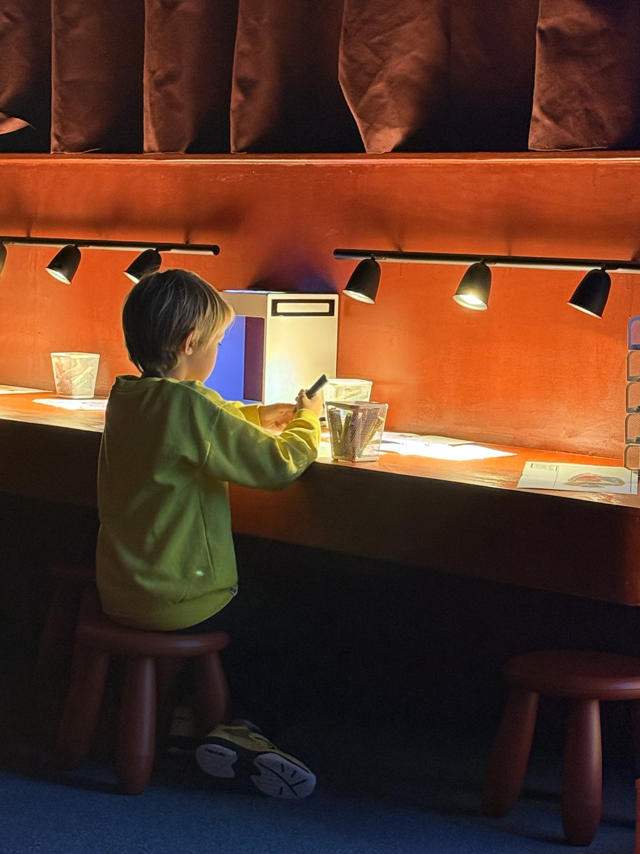 A young boy sitting on a small wooden stool at a booth with a red counter, looking at a smartphone. There are small containers and a paper on the counter, with black track lighting overhead illuminating the area.