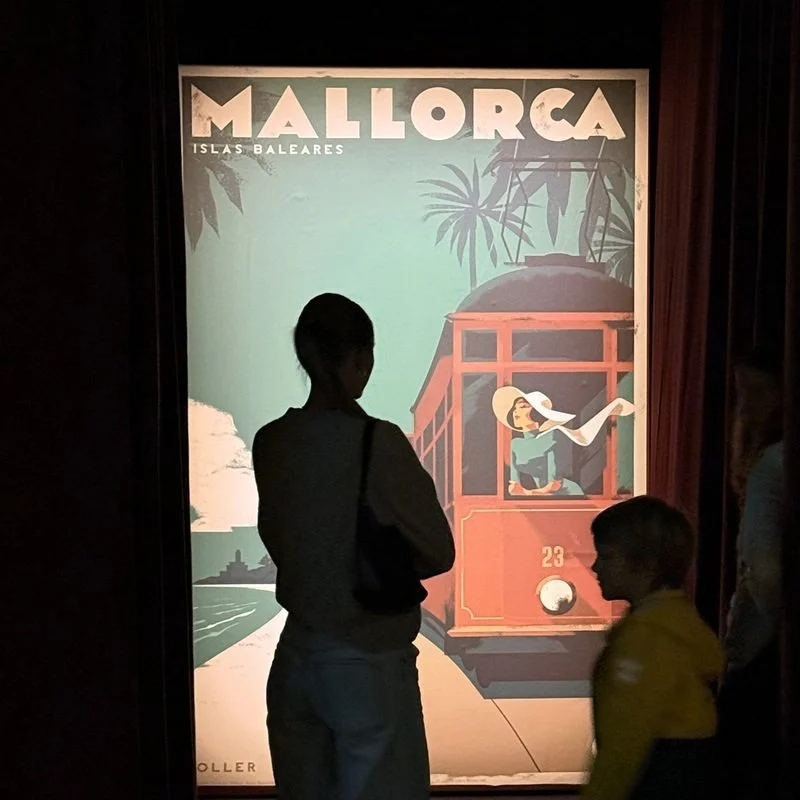 Silhouettes of a woman and a child looking at a vintage travel poster for Mallorca, Islas Baleares, depicting a red tram and a woman in a large hat, with palm trees and a blue sky in the background.