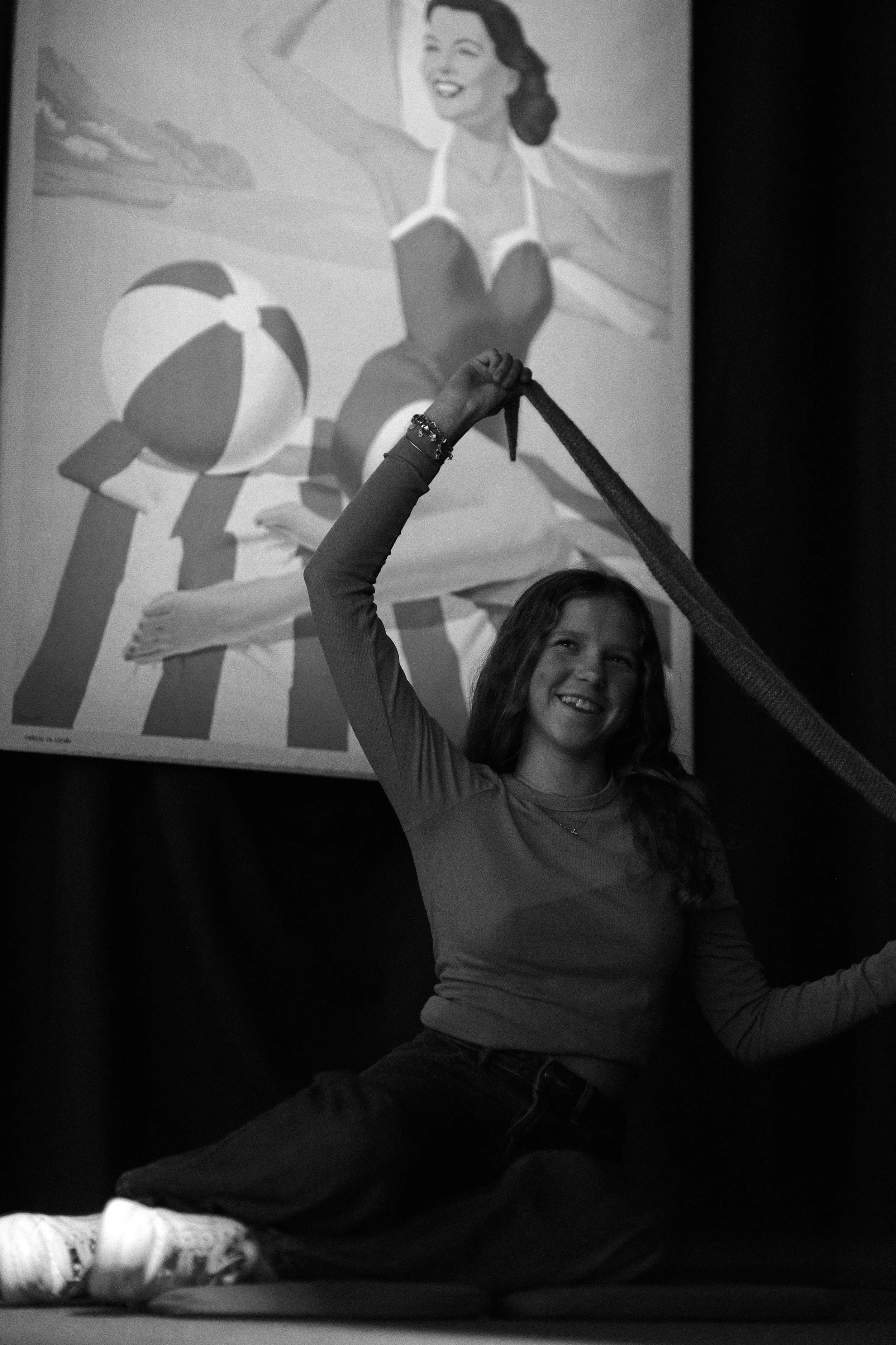 A smiling young woman sitting cross-legged on the floor, holding a rope above her head with one hand, and a vintage-style poster of a woman in a swimsuit posing on a beach in the background.