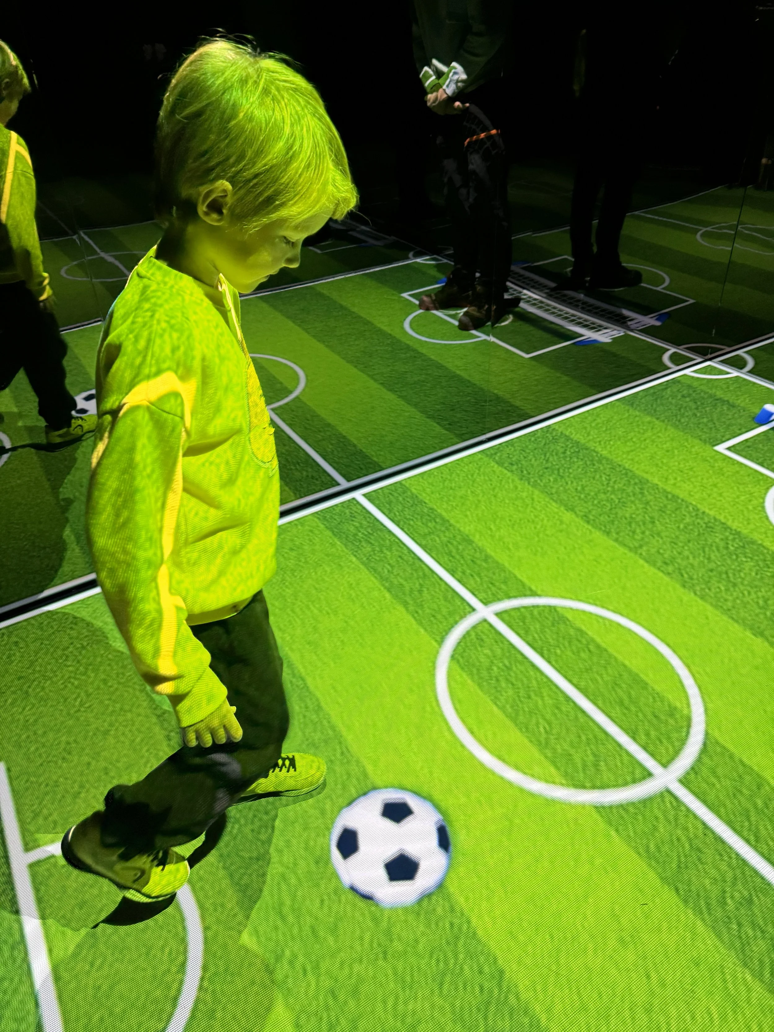 A young boy in a yellow jacket and black pants looking down at a soccer ball on a virtual soccer field with artificial grass, surrounded by mirrors.