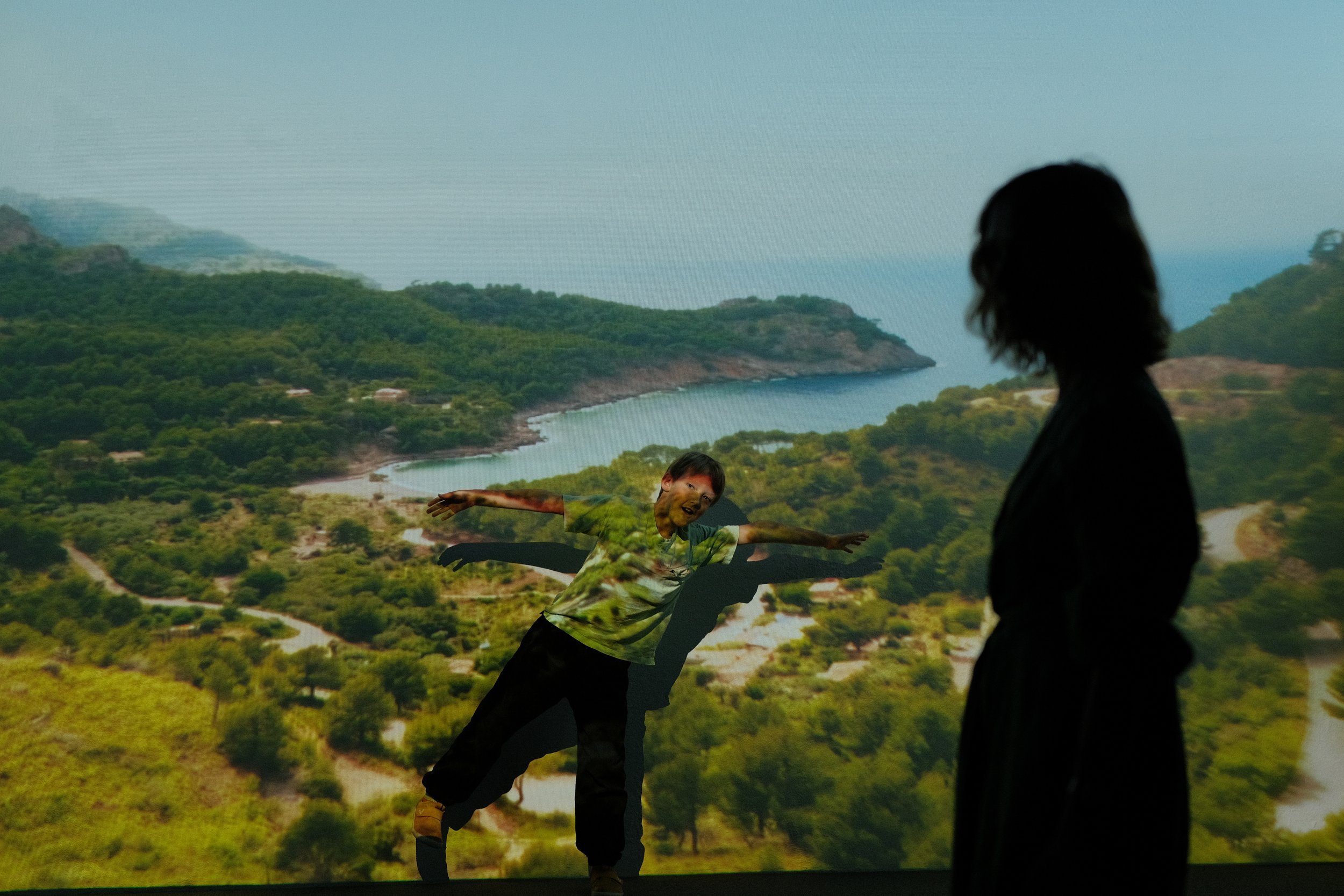 Person standing in front of a digital landscape projection showing a boy with arms outstretched, blended with natural scenery of hills, trees, and a coastline.