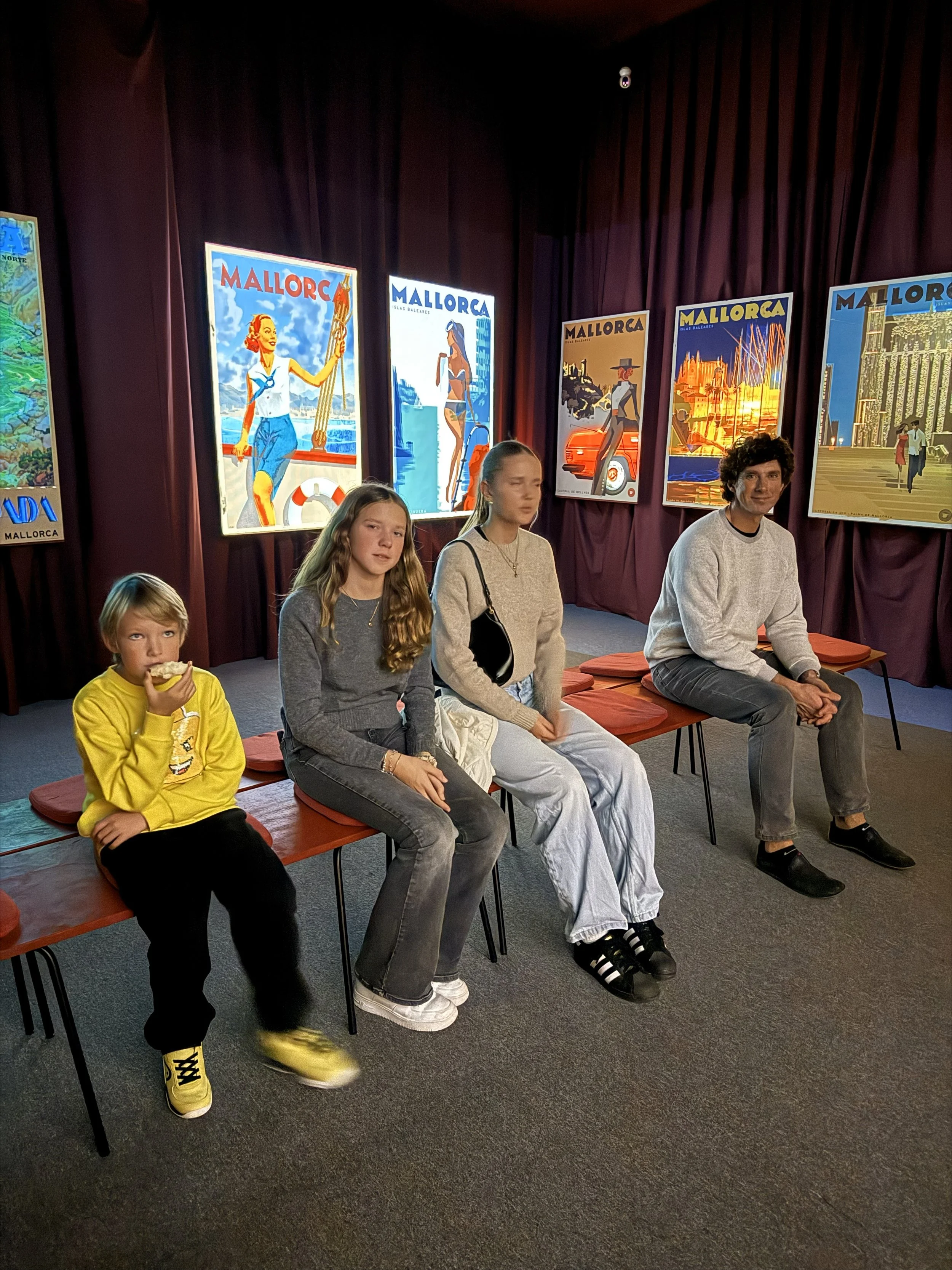 Four people sitting on a bench in front of colorful posters that say "Mallorca" in an indoor setting with purple curtains.