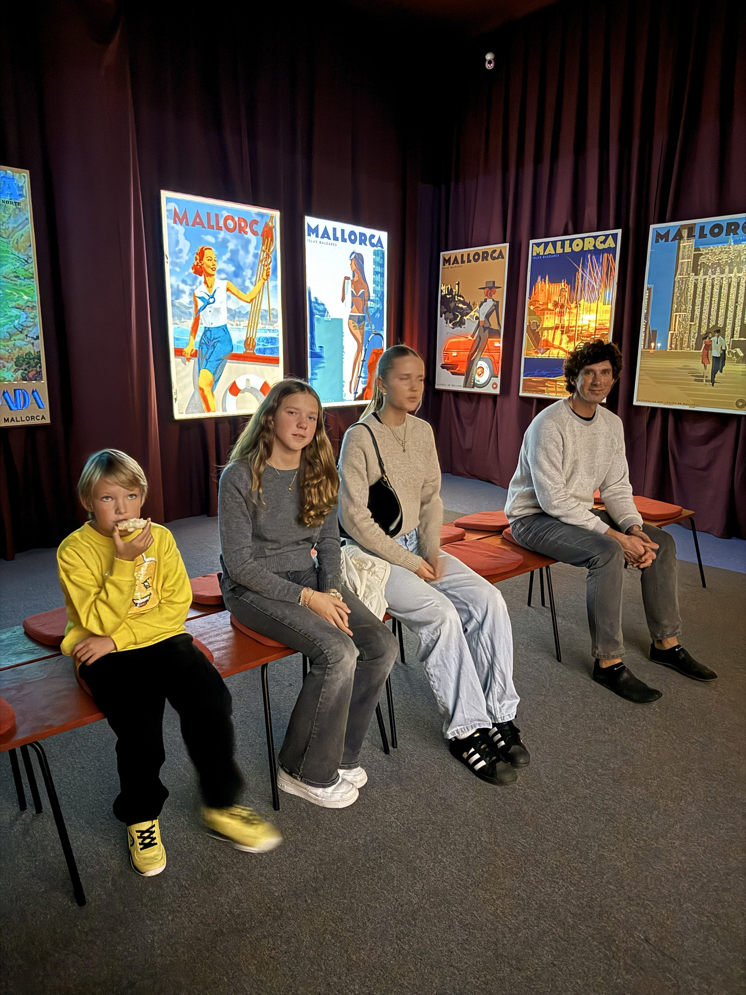 Four people sitting on benches in front of a dark red curtain with colorful Mallorca travel posters on the wall behind them.