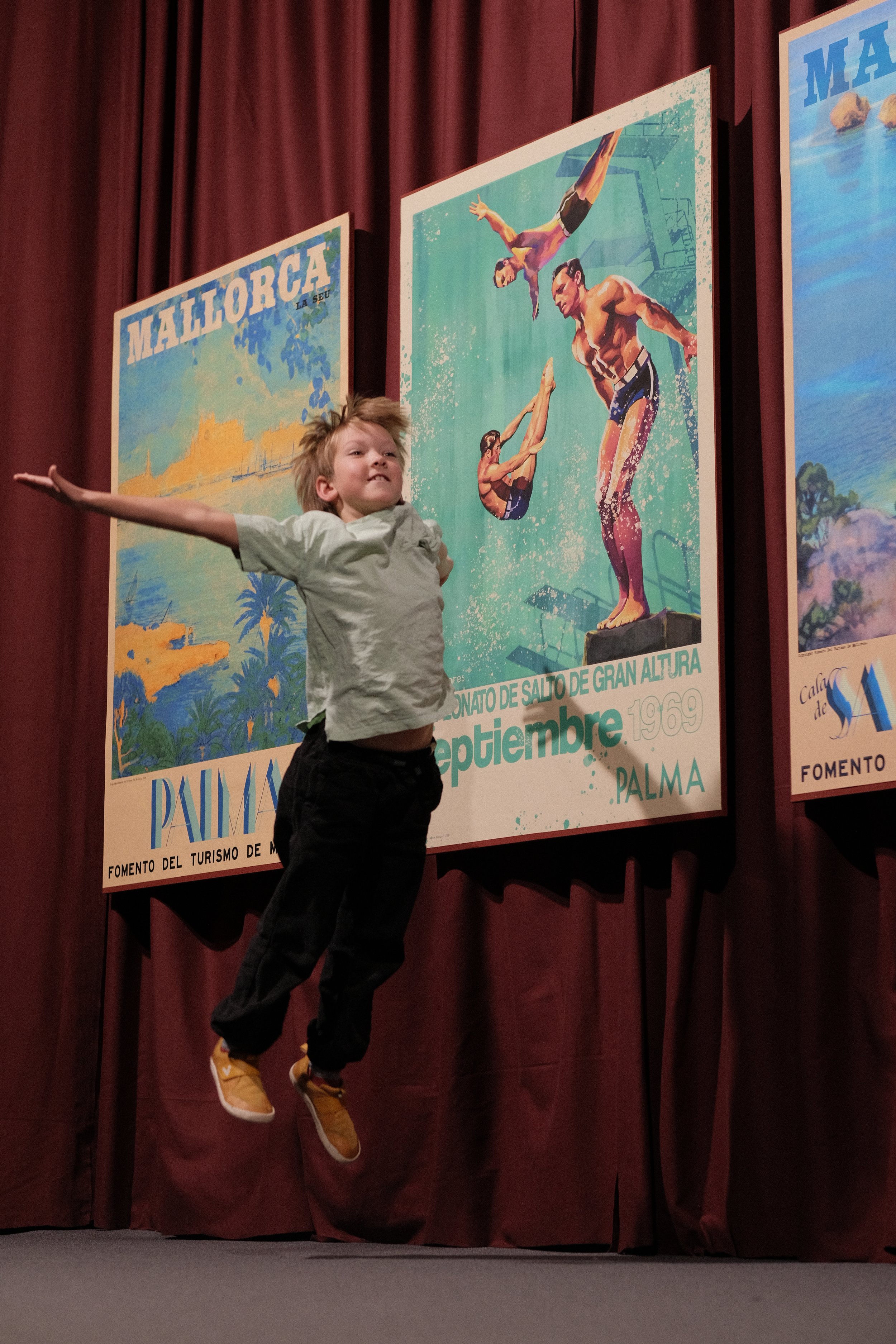 Young boy jumping in front of vintage travel posters from Mallorca and Palma, Spain, displayed on a stage with red curtains.