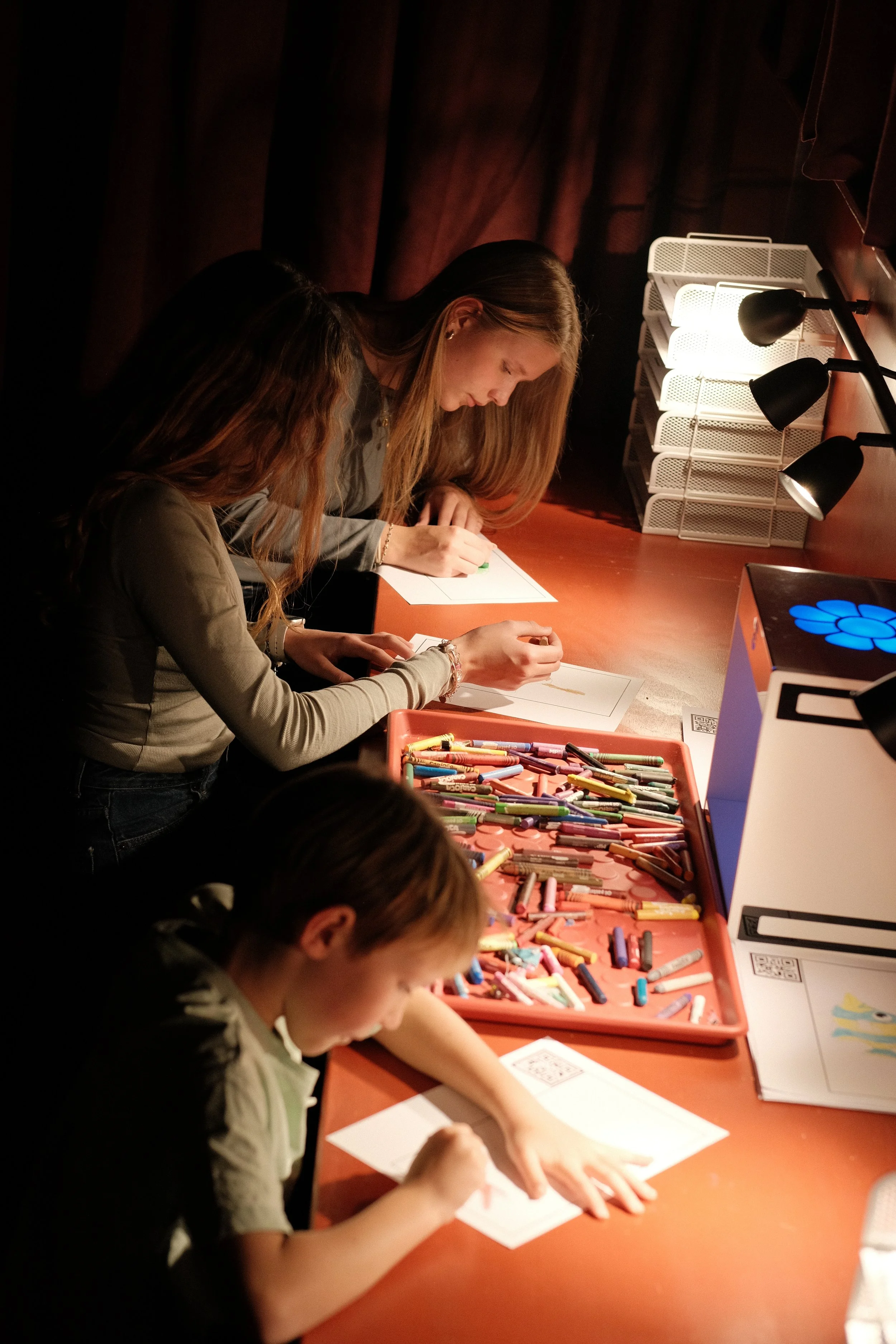 Three girls drawing and coloring with crayons at a table in a dimly lit room.