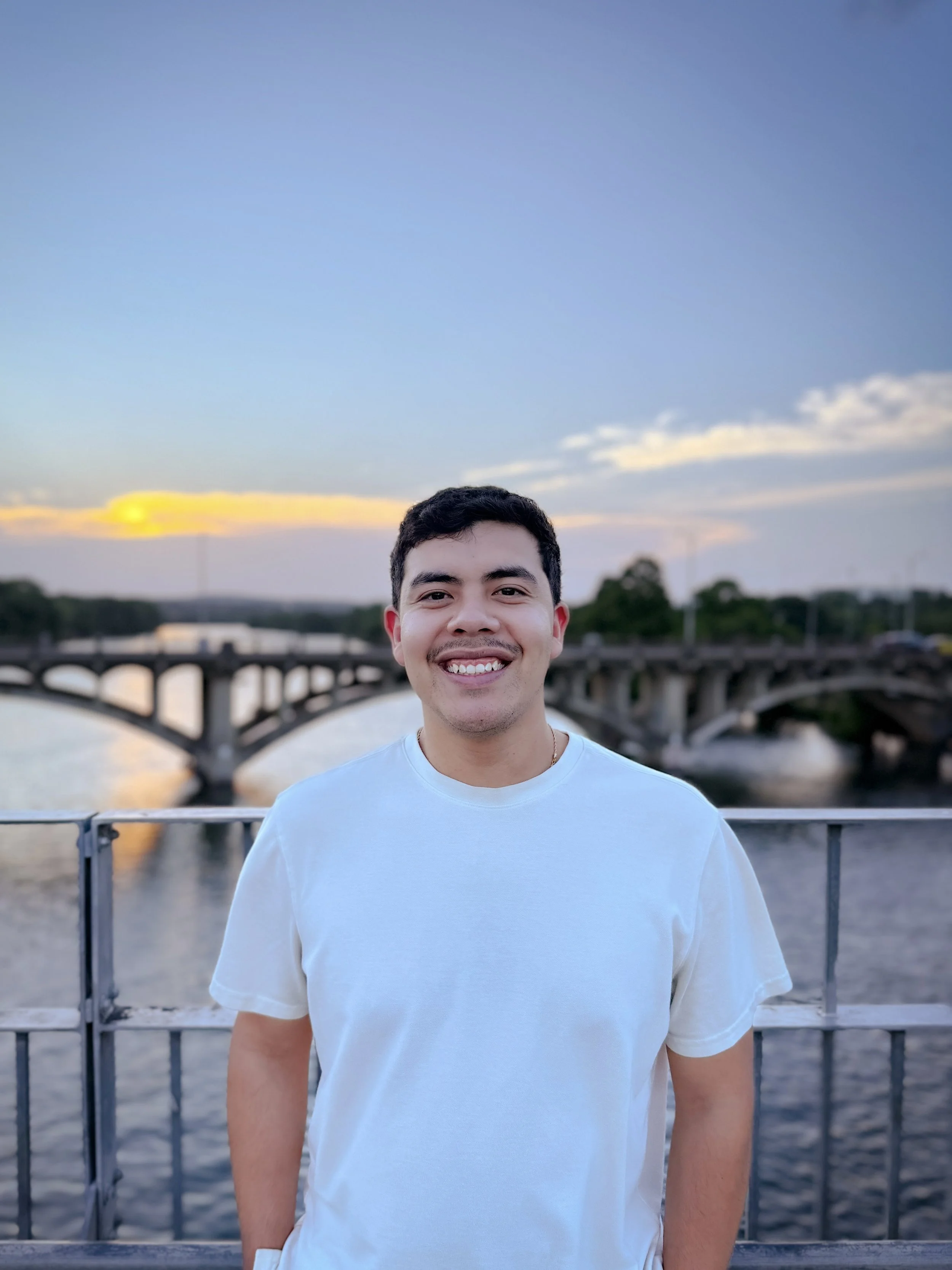 A young man with dark hair and a mustache, wearing a white t-shirt, smiling while standing outdoors near a river at sunset. There is a bridge and trees in the background.