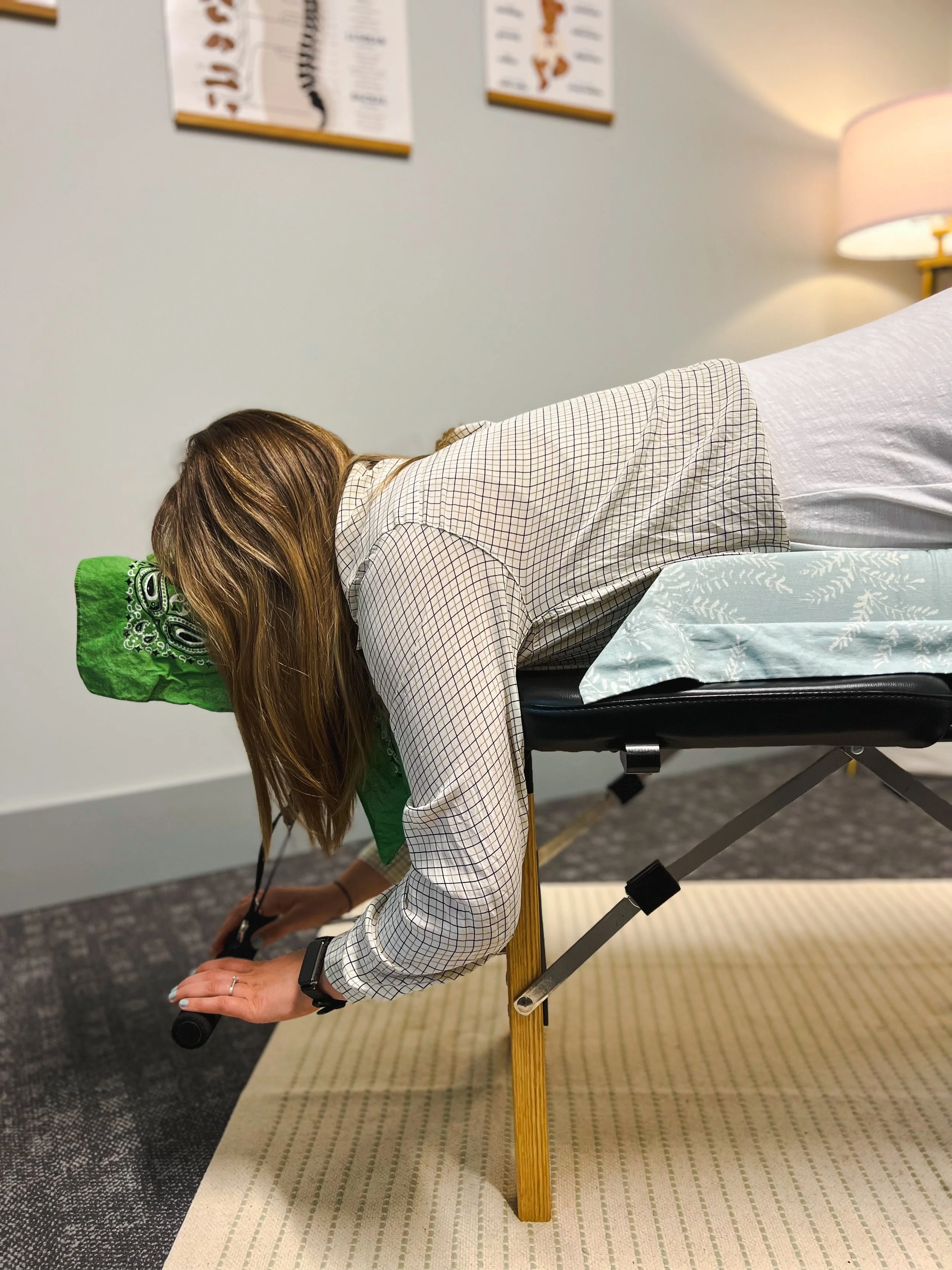 A woman with long brown hair lying face down on a chiropractic table with a green headrest. She is inside a room with beige and dark carpeted floors, white walls, and a lamp in the background.