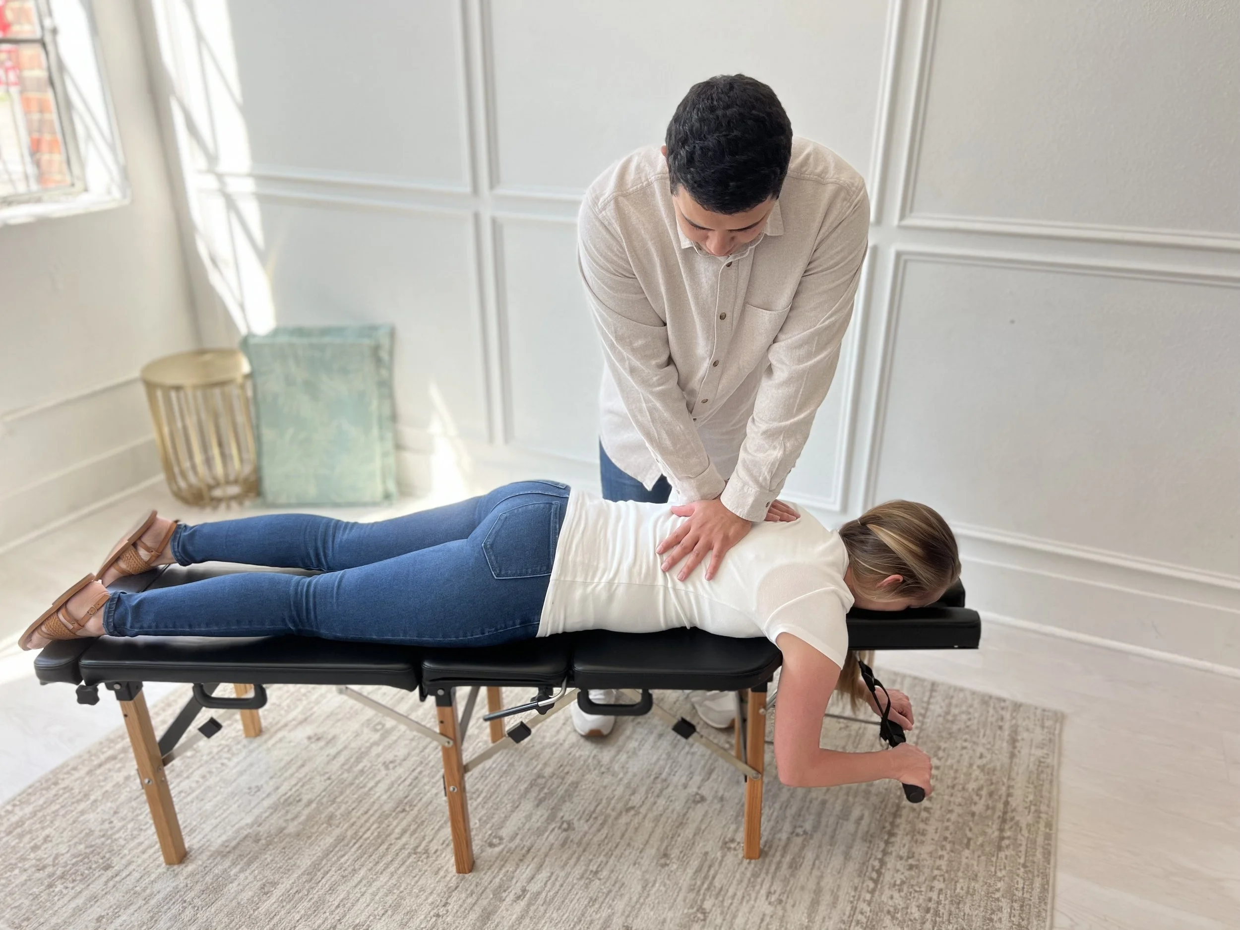 Chiropractic adjustment being performed by a male chiropractor on a female patient lying face down on a chiropractic table, in a well-lit office with white walls, window, and a cushion in the background.