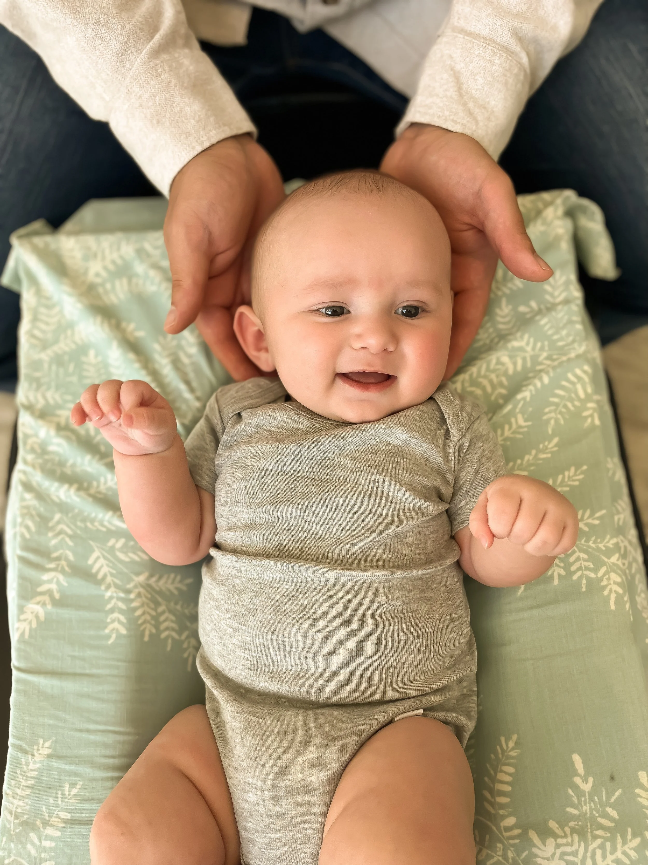 A baby receiving a gentle chiropractic adjustment,  lying on a patterned pillow, being gently held by an adult's hands, smiling and looking happy.