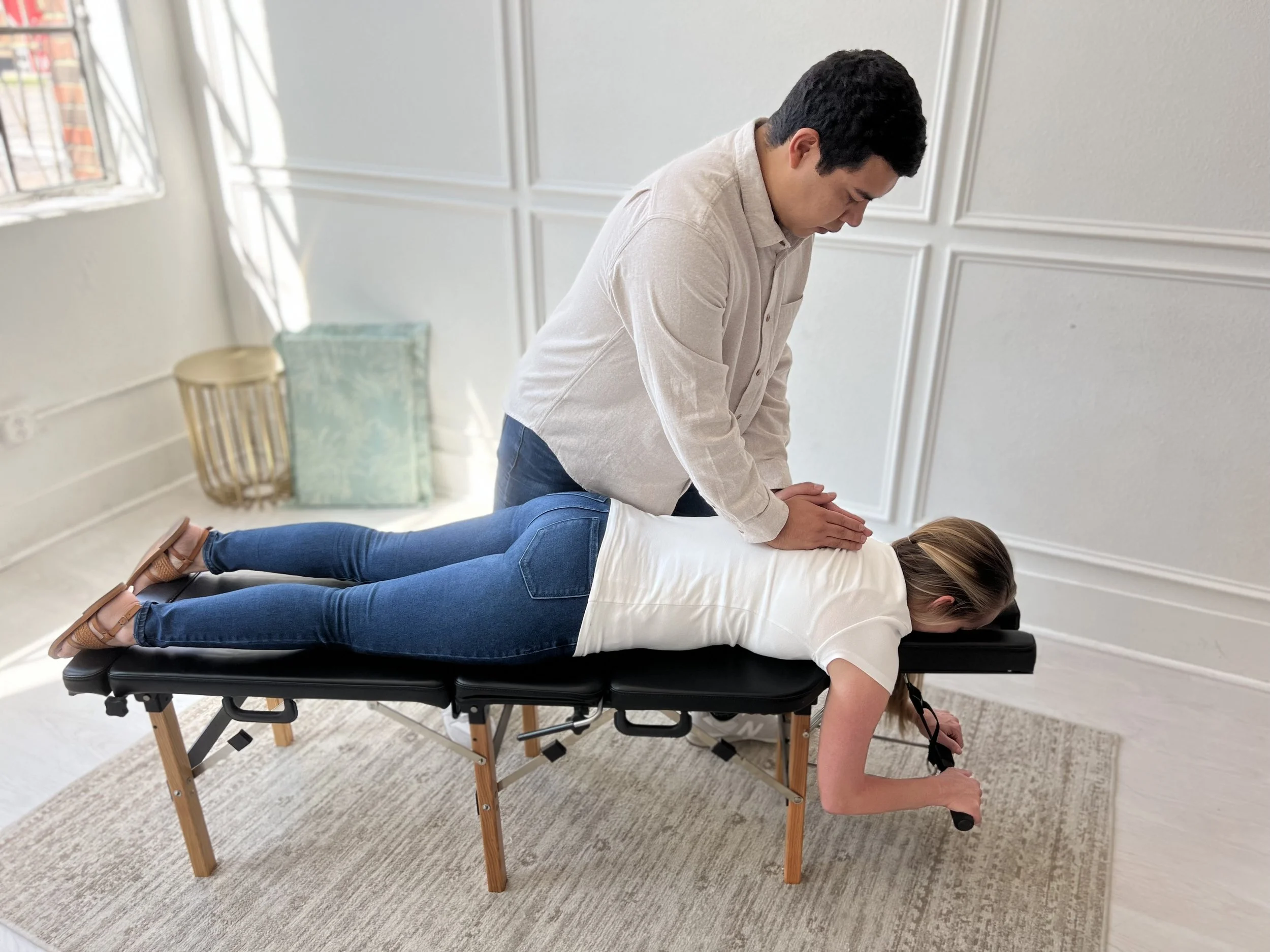 A man providing a chiropractic adjustment to a woman lying face down on a chiropractic table in a bright room.