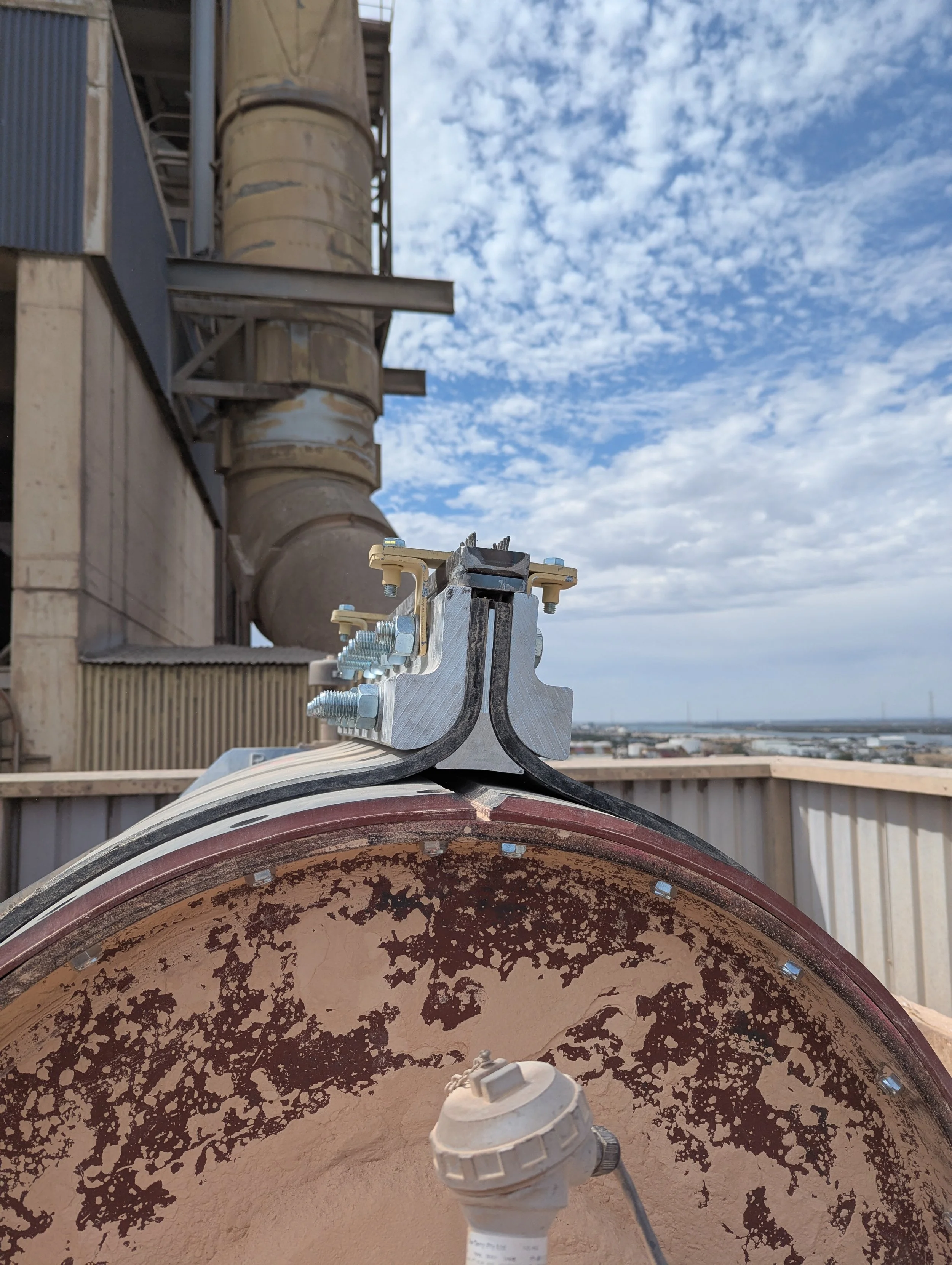 Close-up of a construction or industrial pipe junction with bolts and mounting hardware, set against a background of a metal building, large industrial pipes, and a partly cloudy sky.
