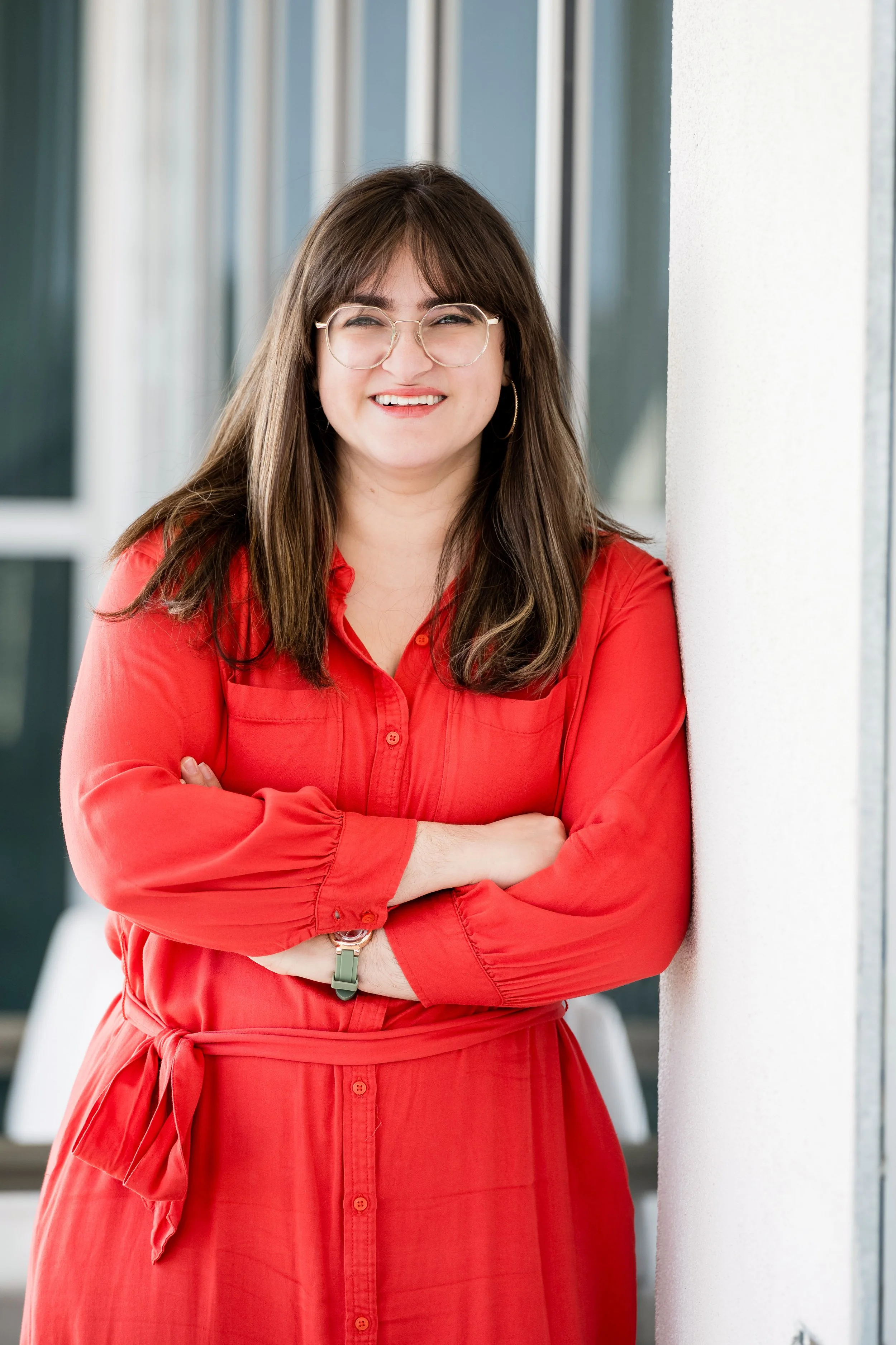 A woman with long brown hair, glasses, and hoop earrings smiling, leaning against a white wall in an indoor setting.