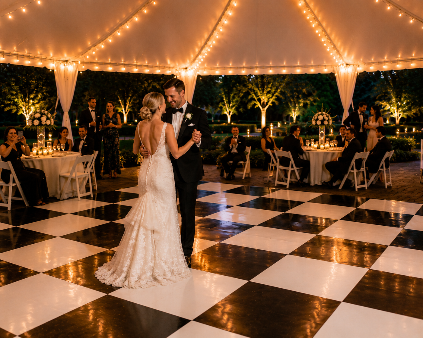 Bride and groom sharing their first dance on a premium black and white checkered dance floor beneath a high peak wedding tent with elegant lighting, white resin chairs, and reception tables at an outdoor wedding reception in Baltimore, Maryland.