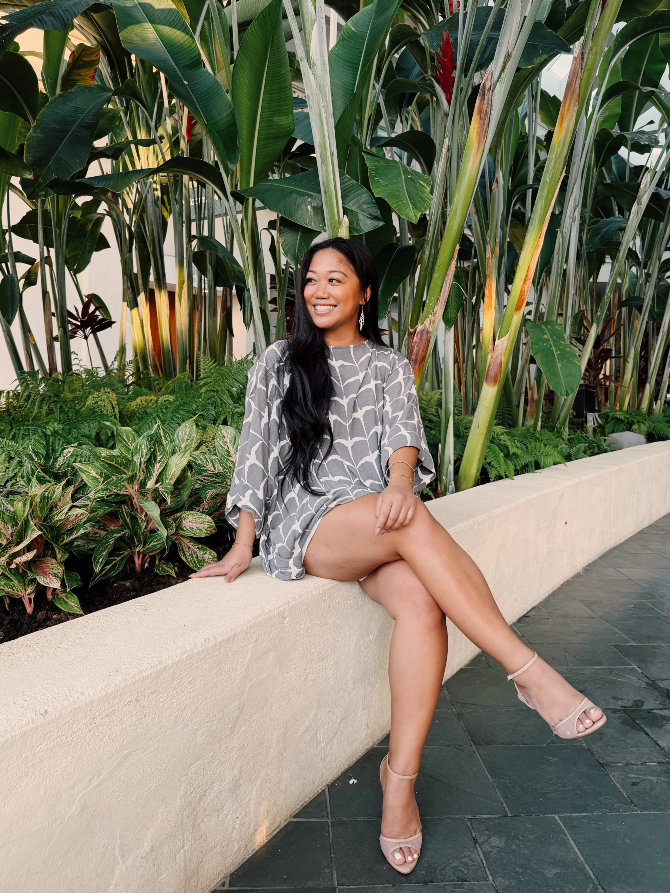 A woman in a patterned dress and nude high heels sitting on a curved beige concrete planter, smiling in front of tall green tropical plants with large leaves.