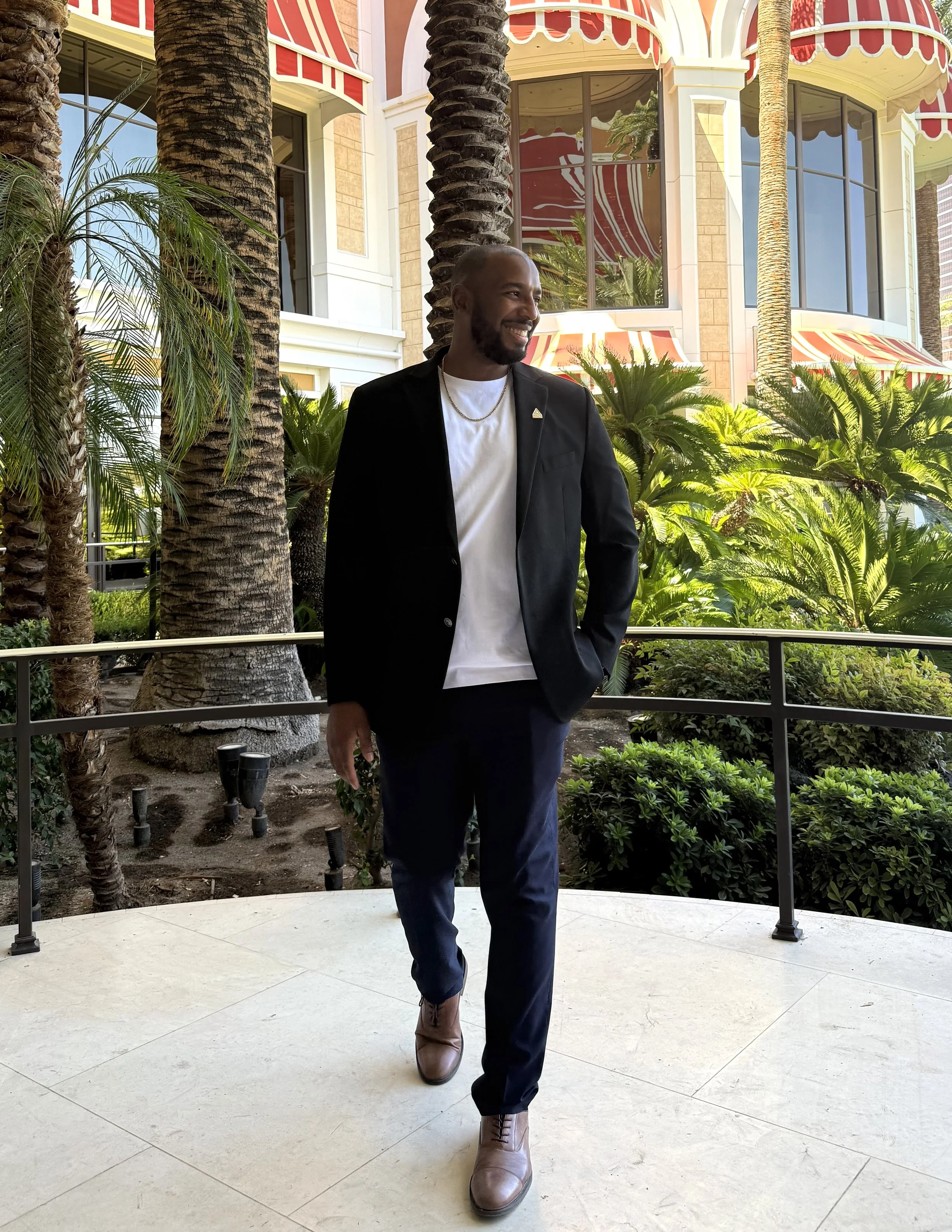 A man wearing a black blazer, white t-shirt, dark blue pants, and brown boots, walking on a light-colored patio with palm trees and lush plants in the background.