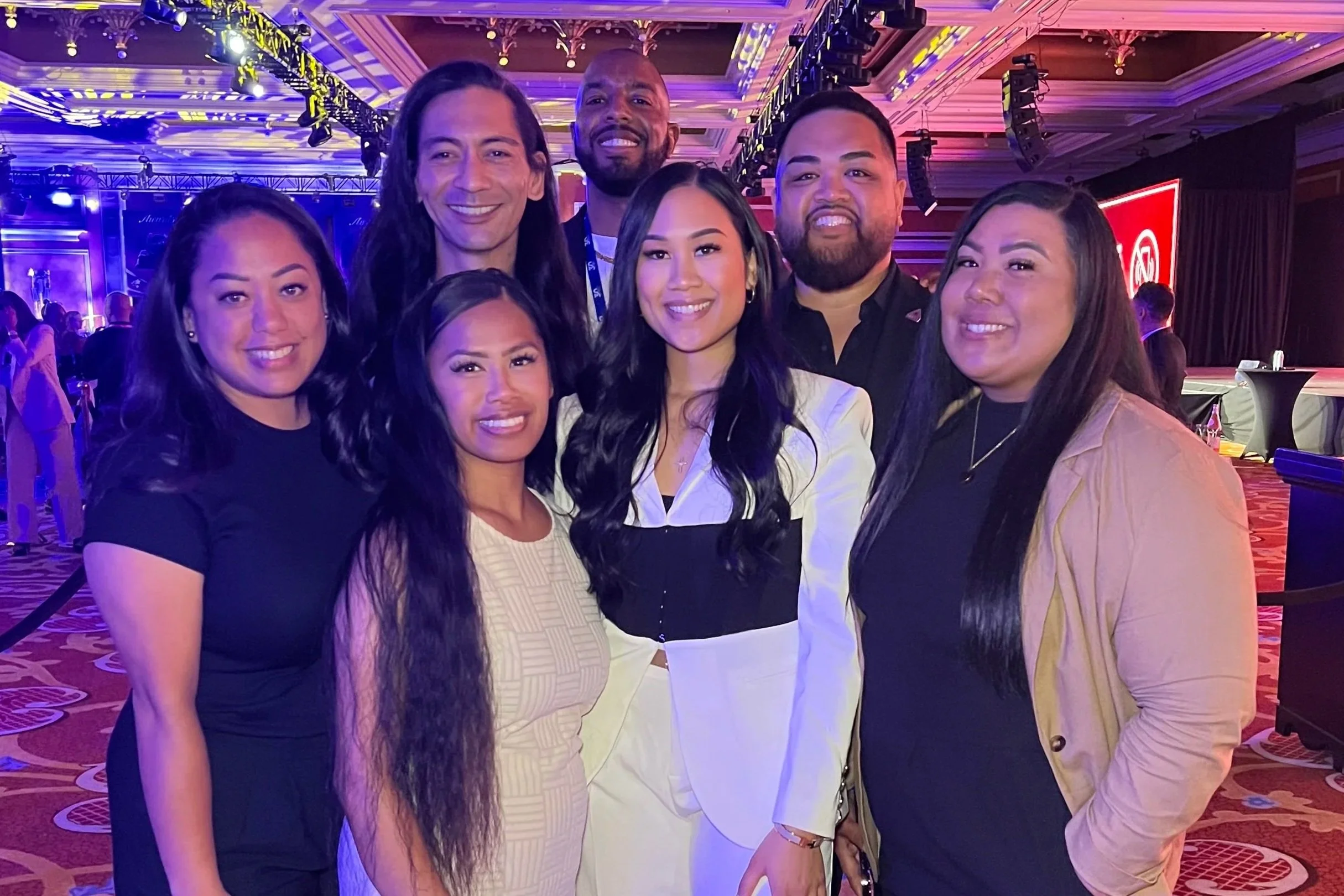 Group of seven diverse people smiling at an indoor event with colorful lighting and stage in the background.