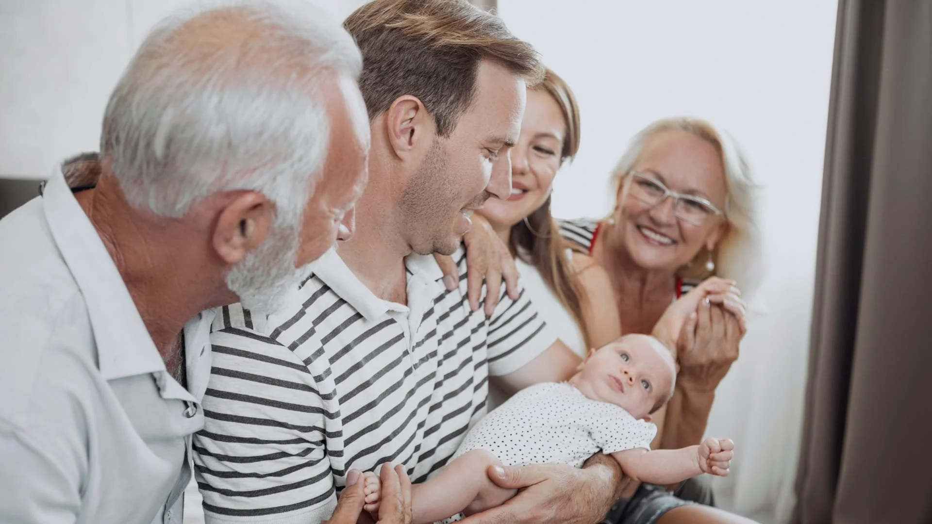 Multi-generational family gathered together, watching something on a television, including a young baby, a middle-aged man, two older women, and an older man, all smiling and engaged.