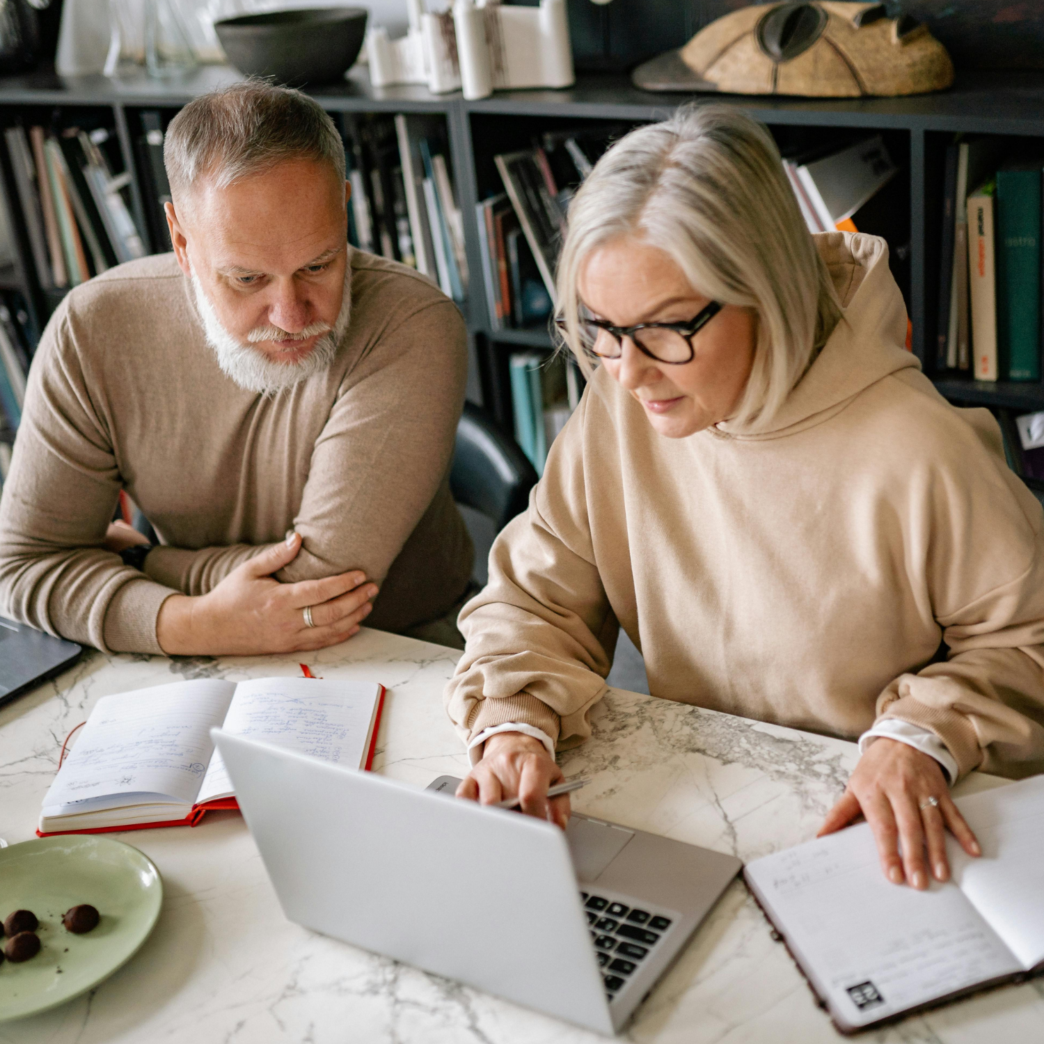 A man and woman sitting at a table looking at a laptop. The woman is pointing at the laptop screen, and the man is watching attentively. There are notebooks, a pen, a plate with chocolates, and a bookshelf in the background.