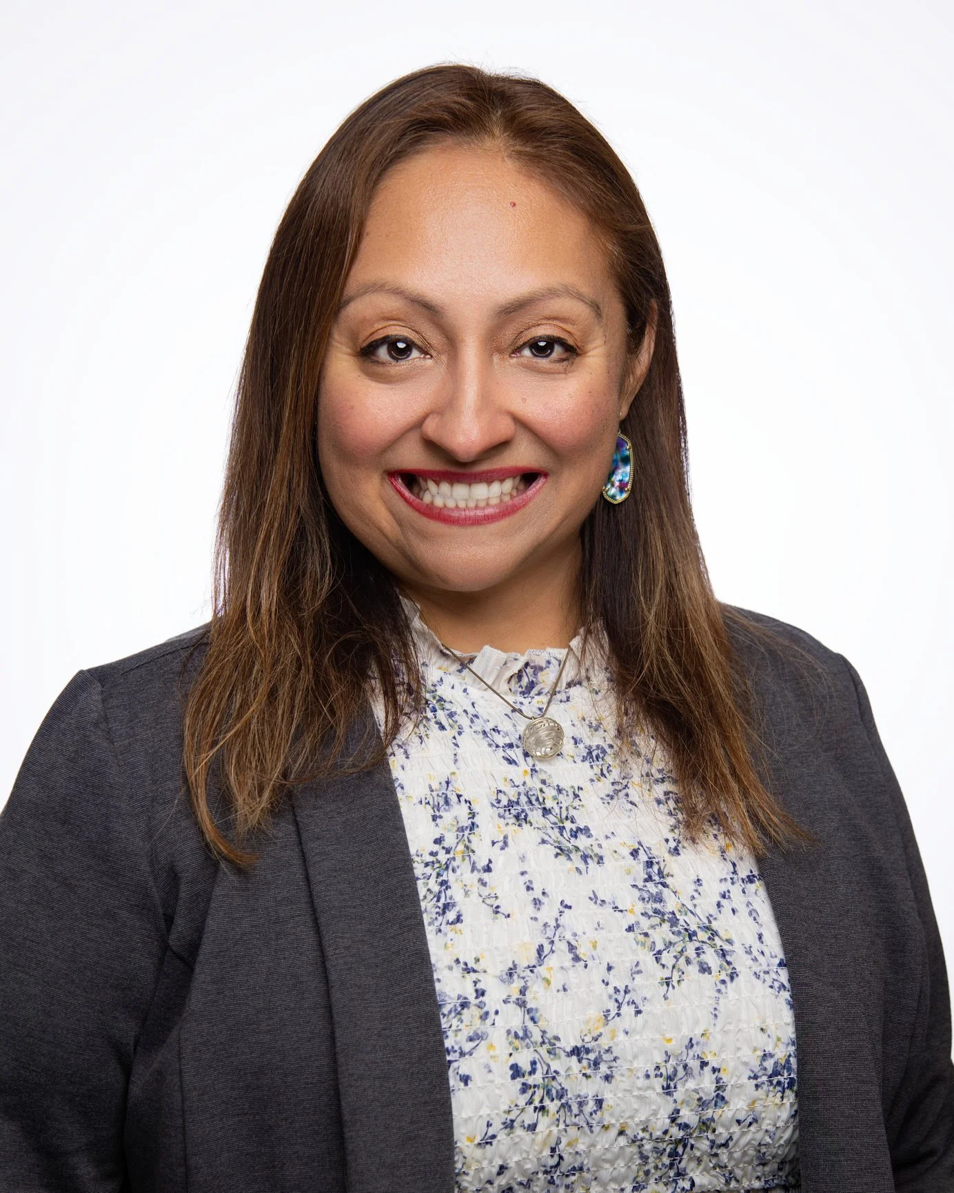 A woman with shoulder-length brown hair, smiling, wearing a dark blazer over a floral blouse, with blue earrings and a necklace, against a plain white background.