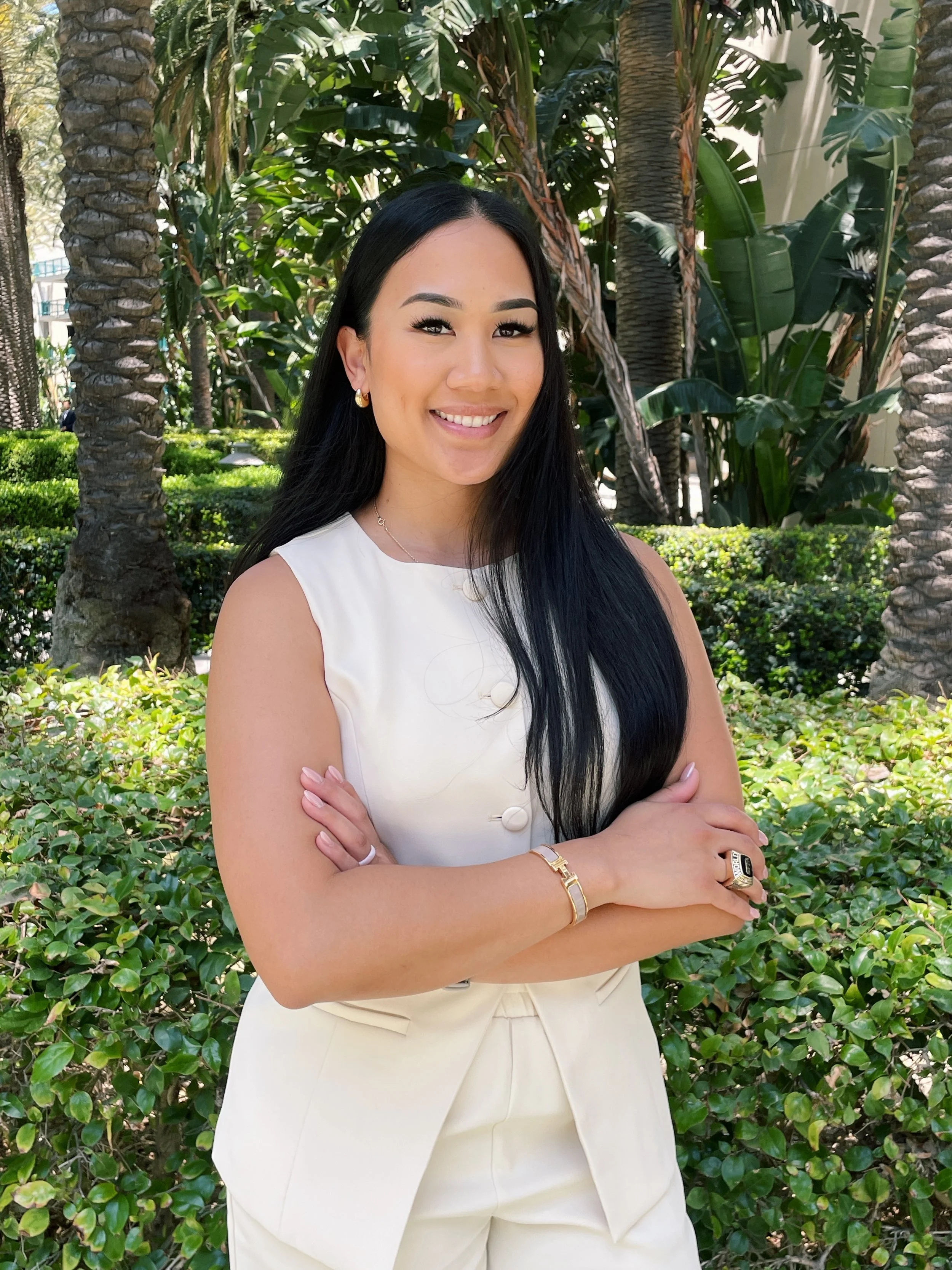 A woman with long black hair wearing a sleeveless white dress stands outdoors with arms crossed, smiling at the camera, surrounded by palm trees and green foliage.