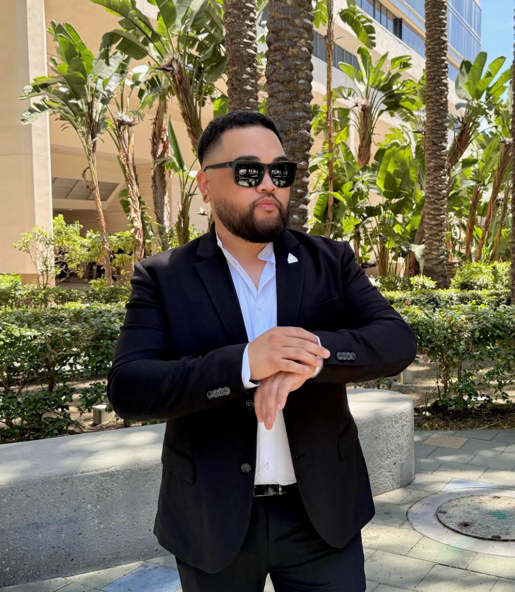 Man in sunglasses and a black suit standing outdoors with palm trees and greenery in the background.