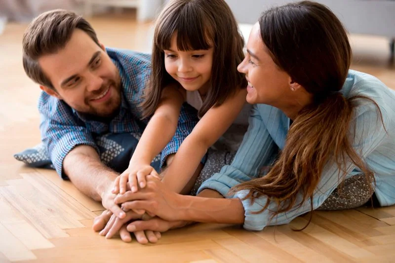 A family playing on the floor together, with a man, woman, and young girl smiling and holding hands.