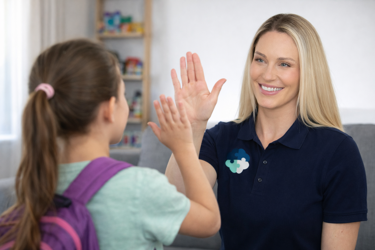 A woman with long blonde hair giving a high five to a young girl with a ponytail, in a cozy room with a shelf in the background.