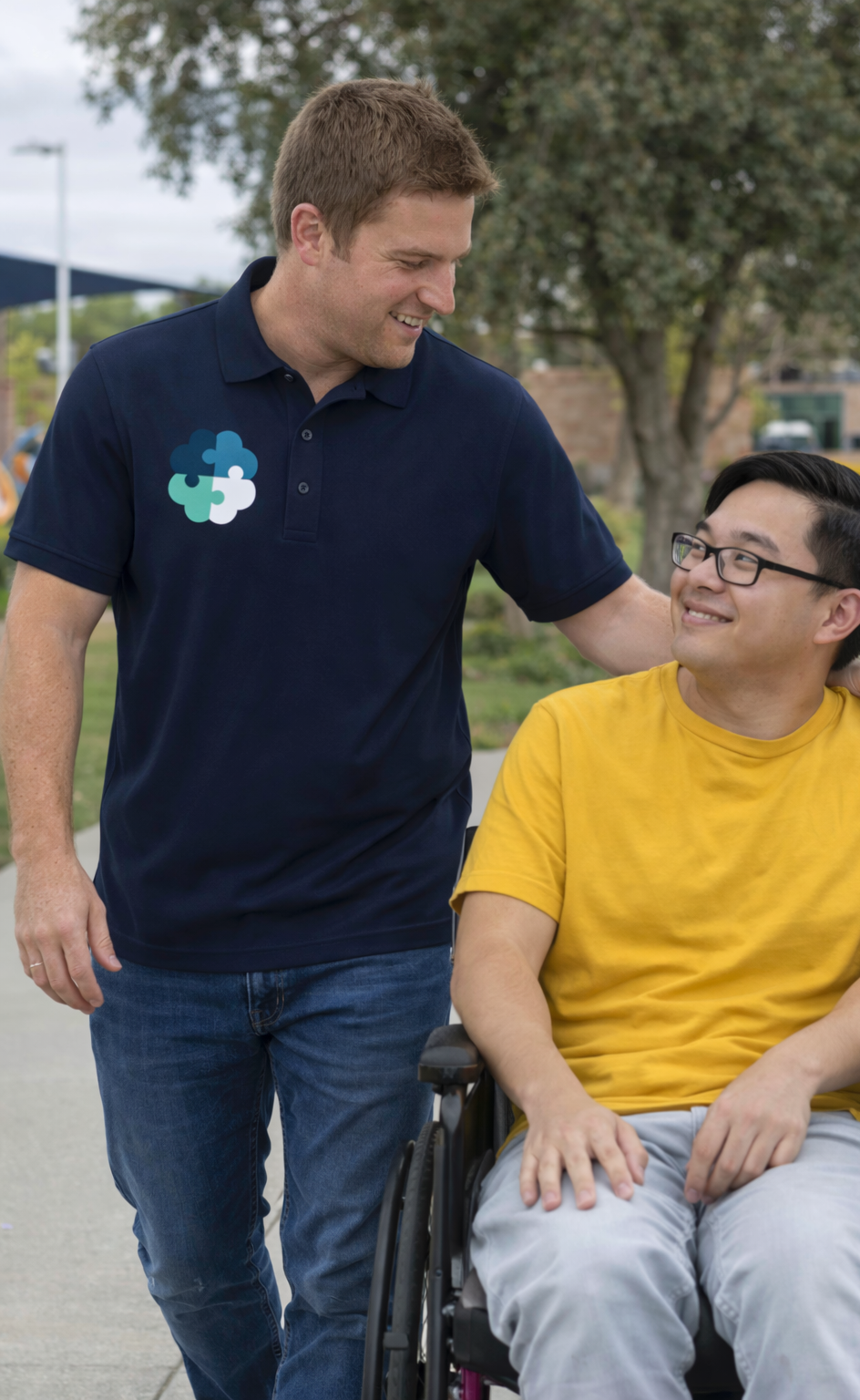 A man in a navy blue polo shirt with a puzzle piece logo is smiling and talking to a man in a yellow T-shirt sitting in a wheelchair.