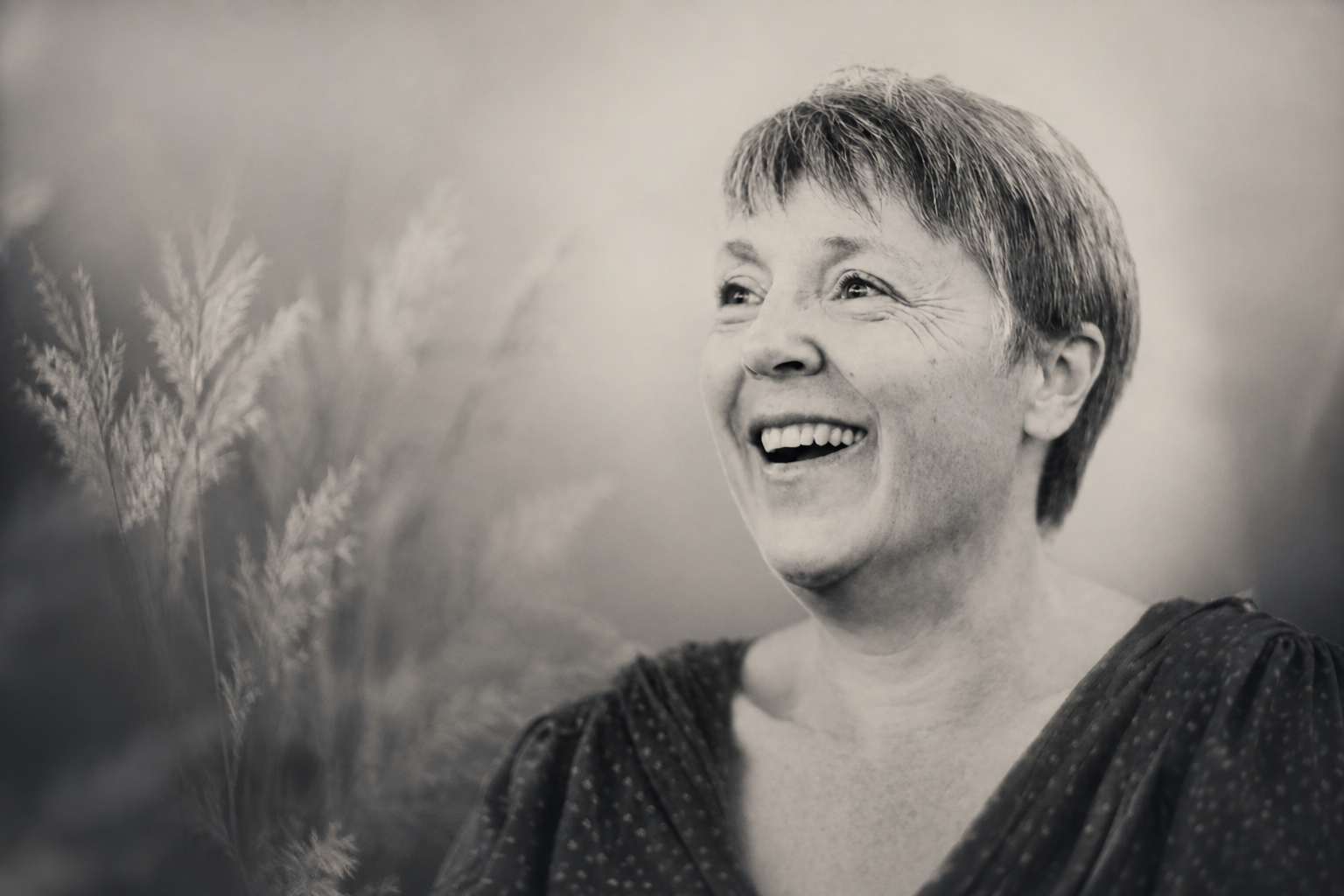 Black and white photograph of a woman smiling and looking into the distance, with some blurred plants in the background.