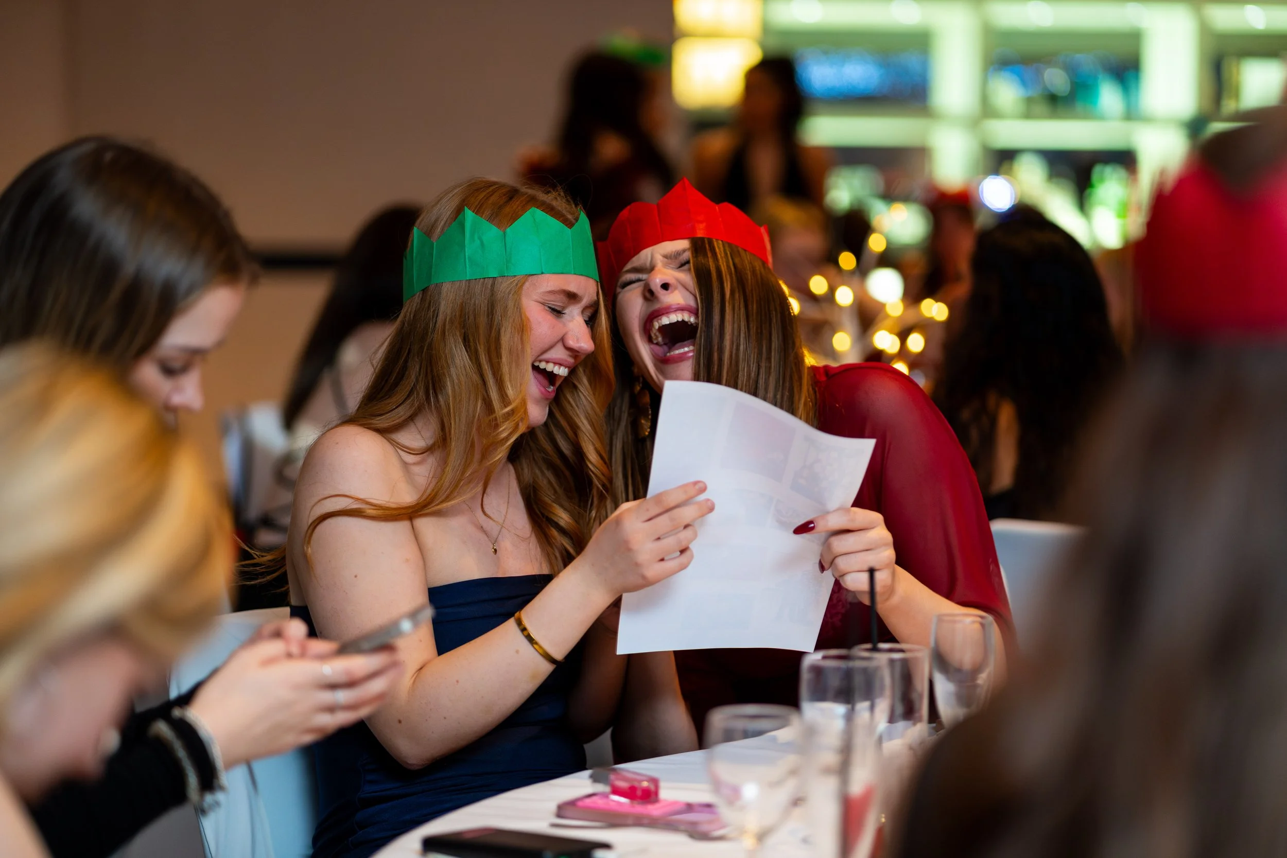 Two women wearing red and green paper crowns laughing and singing together at a party, surrounded by friends and festive decorations.