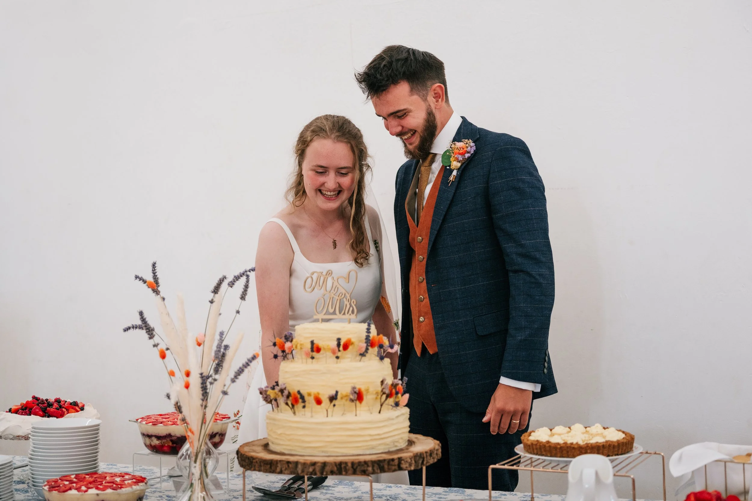 A bride and groom cutting their wedding cake at reception. The bride is wearing a white dress, and the groom is dressed in a dark blue suit with a boutonnière. The cake has a 'Miss & Mrs' topper and is decorated with dried flowers. There are addition
